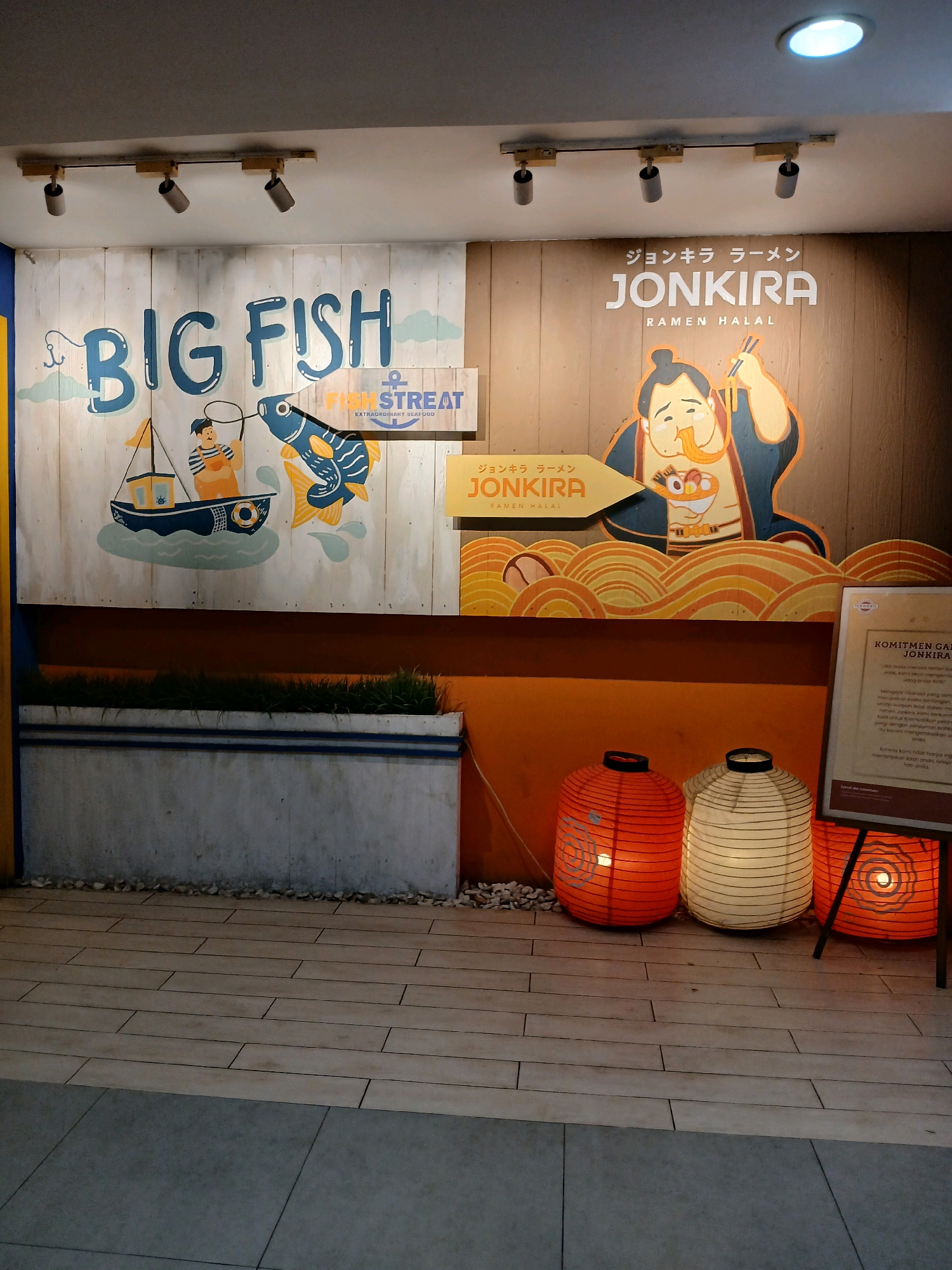 This image depicts the exterior of a restaurant called "JONKIRA RAMEN HALAL" located in South Jakarta, Indonesia. The restaurant features a large mural on its wall. The left side of the mural displays the text "BIG FISH" in large blue letters, along with illustrations of a man fishing from a boat and a large fish. A sign that reads "FISH STREAT EXTRAORDINARY SEAFOOD" is also visible. The right side of the mural features the text "JONKIRA RAMEN HALAL" in white letters against a wooden background, accompanied by an illustration of a man eating ramen. Below the mural, there are three paper lanterns displayed: one orange lantern with a circular pattern, one white lantern with horizontal stripes, and another orange lantern with a circular pattern. A concrete planter with grass is situated below the left side of the mural. A sign with text in Indonesian is also visible to the right of the lanterns. The flooring appears to be made of wood tiles. The scene is illuminated by artificial lighting, including track lighting on the ceiling and a circular ceiling light.