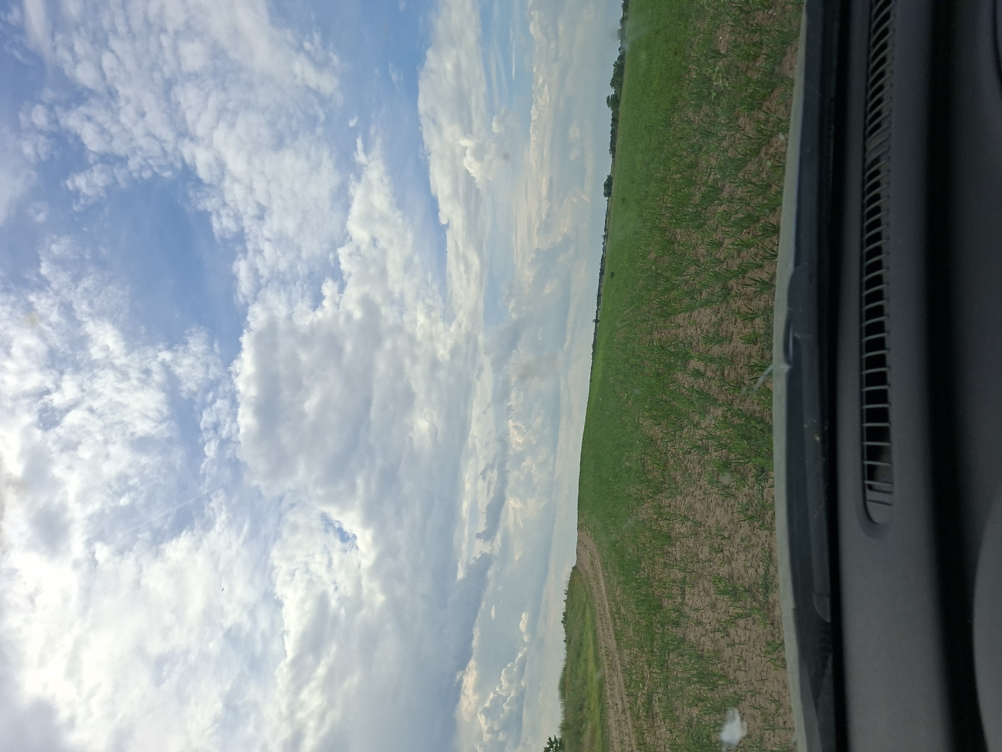 The image, likely taken from inside a vehicle, shows a view of the sky and a field. The sky is predominantly blue with a large number of white clouds. A field is seen on the right side of the frame; green plants are sparsely covering the brown earth. The right-most edge of the frame includes a portion of the car interior, specifically a gray panel with ventilation slots. The photograph seems to be taken at an angle, tilting to the left. Based on the prompt, the location is the city of Pobeda, Bulgaria.