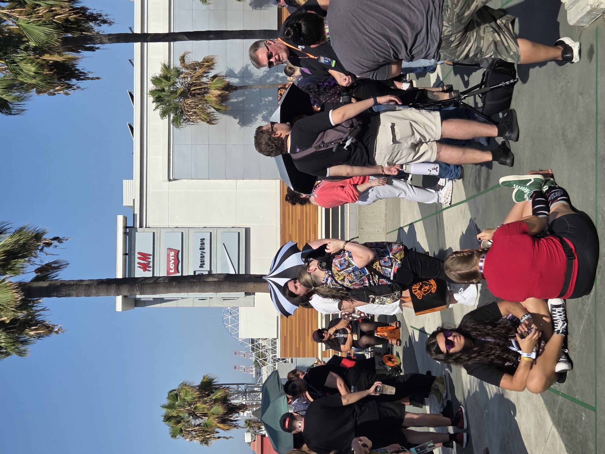 In Long Beach, California, under a clear blue sky, a group of people gathers outdoors in front of a modern building. The building features prominent signage for H&M, Levi's, and Factory Store. Palm trees, characteristic of the Southern California landscape, frame the scene. Several individuals are standing, some holding umbrellas, and others are seated on the ground. One person is taking a photograph, while two men engage in conversation. In the background, a Ferris wheel is visible, suggesting the location might be near an amusement park or waterfront attraction. The ground appears to be paved, possibly a plaza or walkway. The overall atmosphere suggests a casual outdoor event or gathering.