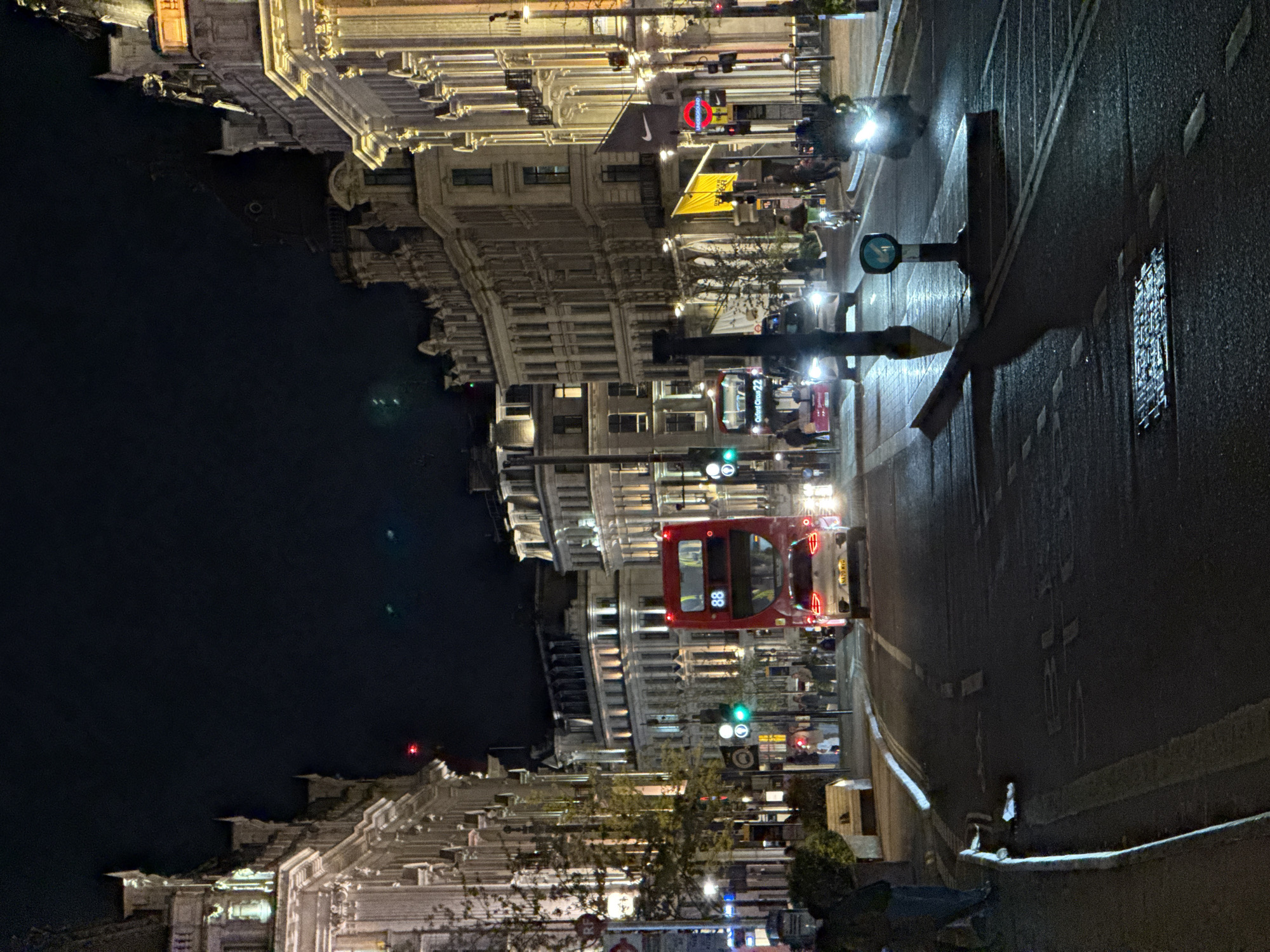 The image shows a nighttime street scene in London, United Kingdom. A red double-decker bus with the number 88 is traveling down the street, which has wet asphalt reflecting the streetlights. The street is lined with buildings that have ornate facades, lit up by streetlights and the lights from within the buildings. Some pedestrians are visible along the sidewalk. A circular underground sign is also visible.