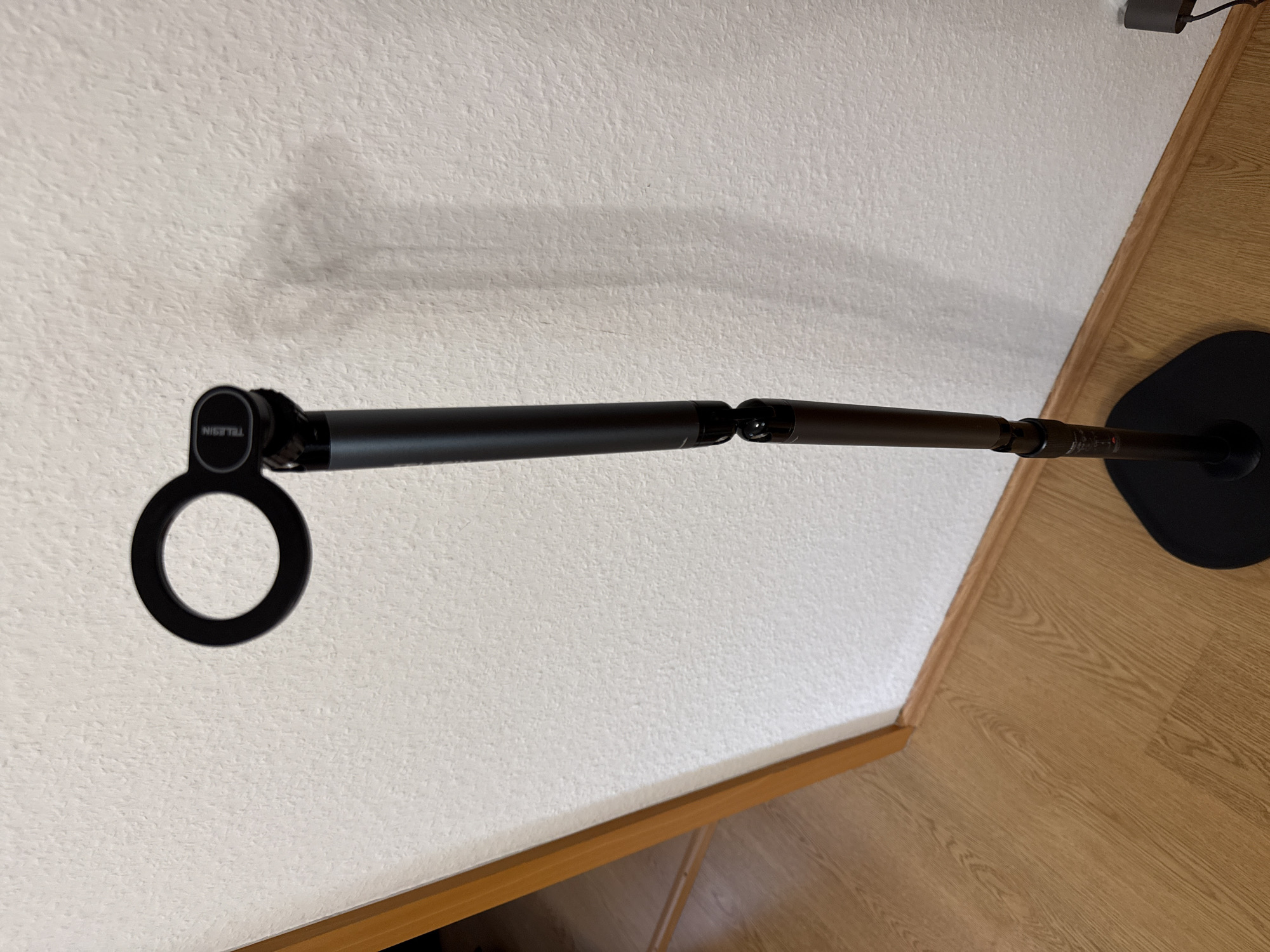 This media file shows a black articulating arm attached to a circular black base, which is resting on a wooden floor. The arm extends horizontally across the frame, reaching towards a white textured wall. The arm itself appears to be made of dark metal and has visible joints allowing for adjustability. On the left side of the arm, a circular attachment with the word "TELESIN" is visible. The lighting suggests an indoor environment, possibly during the daytime, as there are no distinct shadows indicating artificial lighting. The scene is static, with no people or active events depicted. The overall impression is a functional object, likely a mount or stand, positioned in a domestic or office setting. The floor has a wood grain pattern, and the wall has a textured finish.