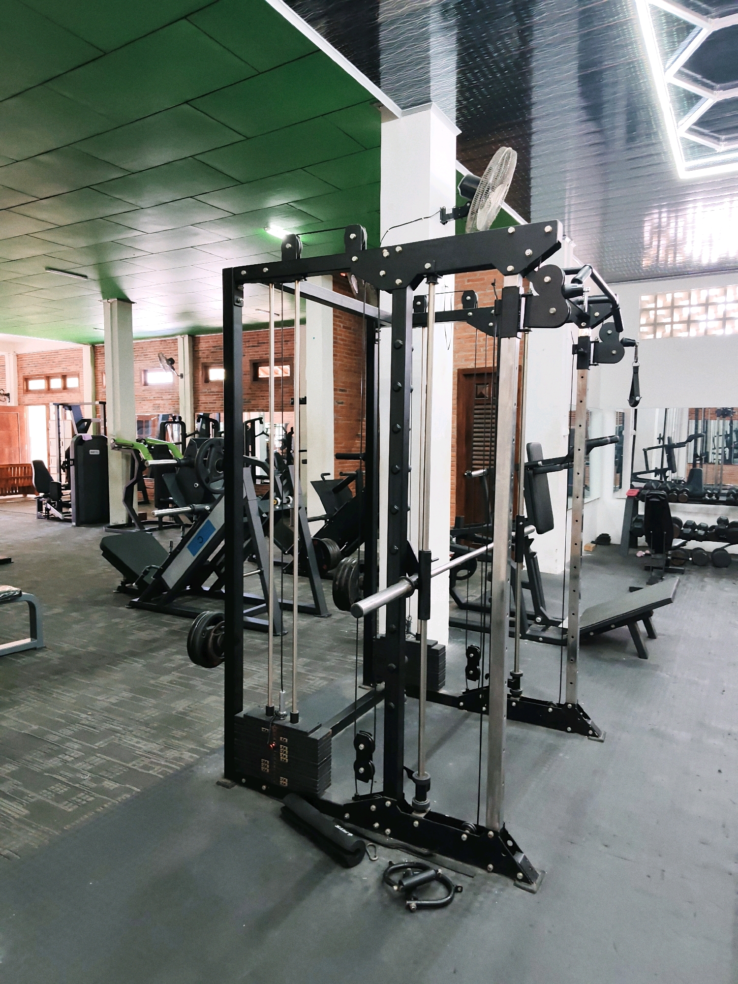 This is a medium-angle shot of a gym in Pancur, Indonesia. The gym has a green ceiling with reflective panels. The walls are made of red brick. In the foreground is a multi-gym machine with black metal framing and weight stacks. To the left of the multi-gym is a leg press machine. In the background, there are other weight machines, dumbbells, and benches. The floor is covered with grey rubber mats. A ceiling fan is visible on the wall in the background.