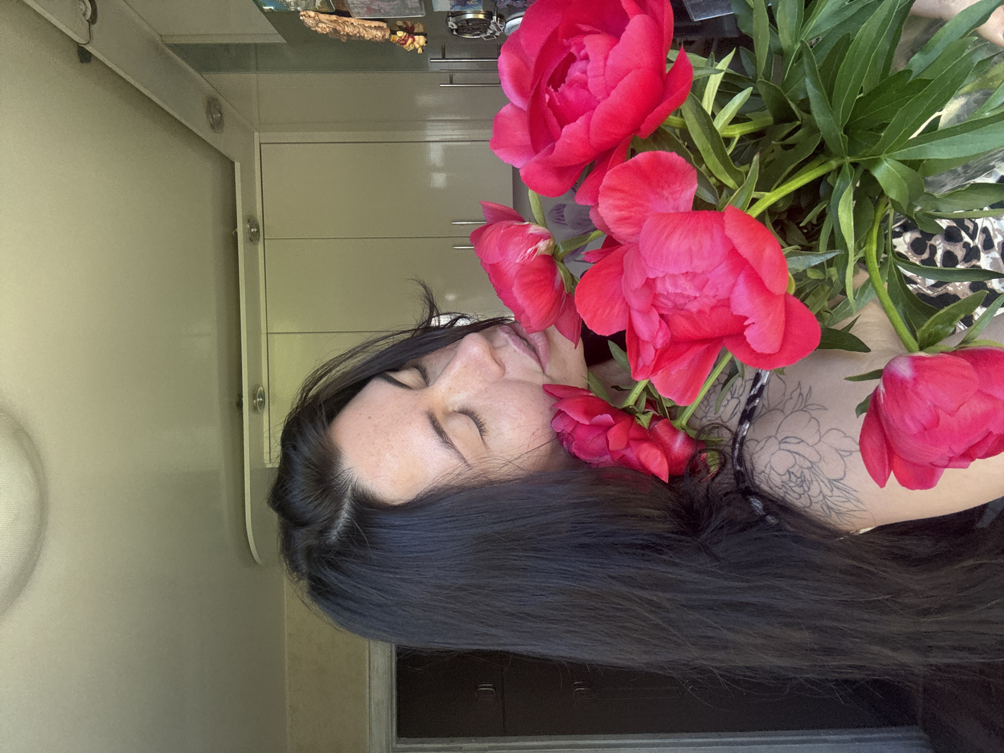 The image shows an indoor scene in Chaikovsky, Russia, featuring an adult female subject smelling a large bouquet of vibrant magenta-red peonies. Her eyes are closed, and her face is gently pressed into the flowers. She has long, dark hair, and freckles are visible on her forehead. An uncolored outline tattoo, possibly floral, is visible on her left upper arm. She is wearing a patterned garment. The background consists of a domestic interior, likely a kitchen, featuring light-colored cabinetry with chrome handles, recessed ceiling lights, and smaller task lights below a ceiling bulkhead. A dark brown door is present on the left, and various small decorative items are partially visible on the far right.