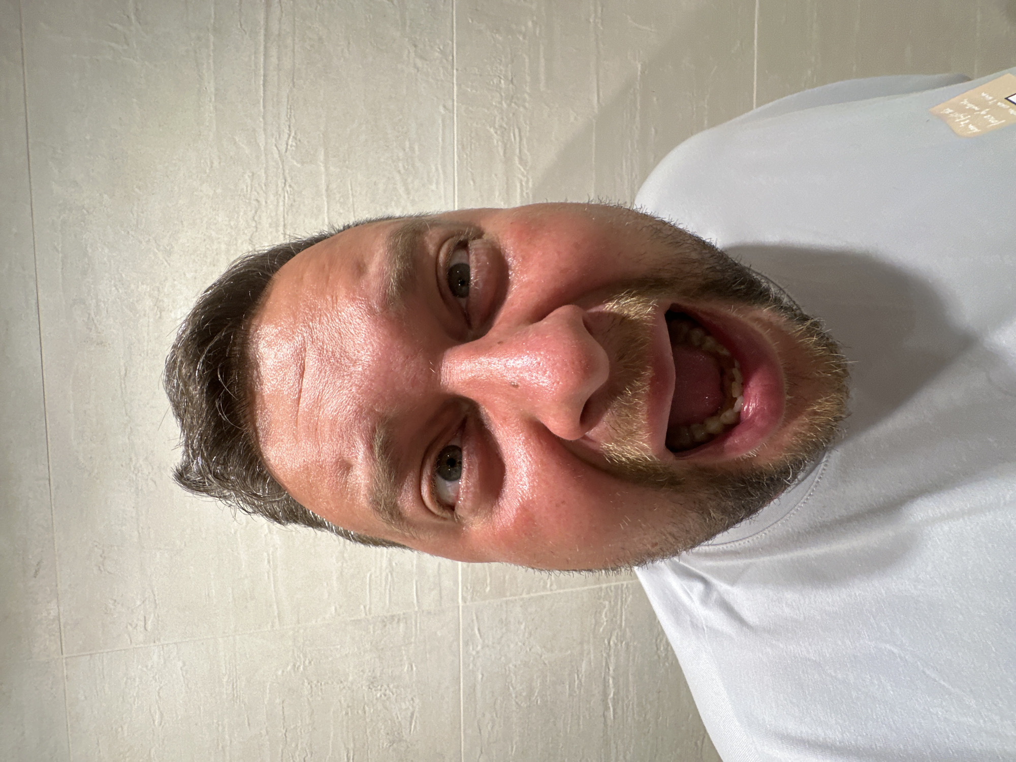 A close-up, eye-level, full-face portrait shows a Caucasian man with light brown hair and stubble. He is wearing a light blue t-shirt and is positioned in front of a tiled wall. The man is making a facial expression with his mouth open wide and his eyes looking to the side. The tiles on the wall are a neutral color and are laid out in a horizontal pattern. The lighting appears to be artificial and is casting shadows on the man's face. The overall scene suggests a casual, indoor setting, possibly a bathroom or a similar tiled room. There are no other people or objects of note in the image.