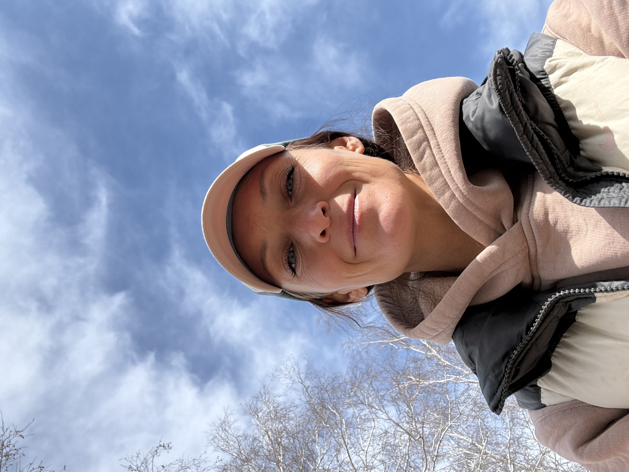 A woman wearing a light pink hooded sweatshirt and a light-colored baseball cap is looking down at the camera with a slight smile. The sky behind her is blue with white clouds. Bare tree branches are visible at the bottom of the frame. The location cannot be determined to be Ulyanovsk, Russia from the information provided in this image.
