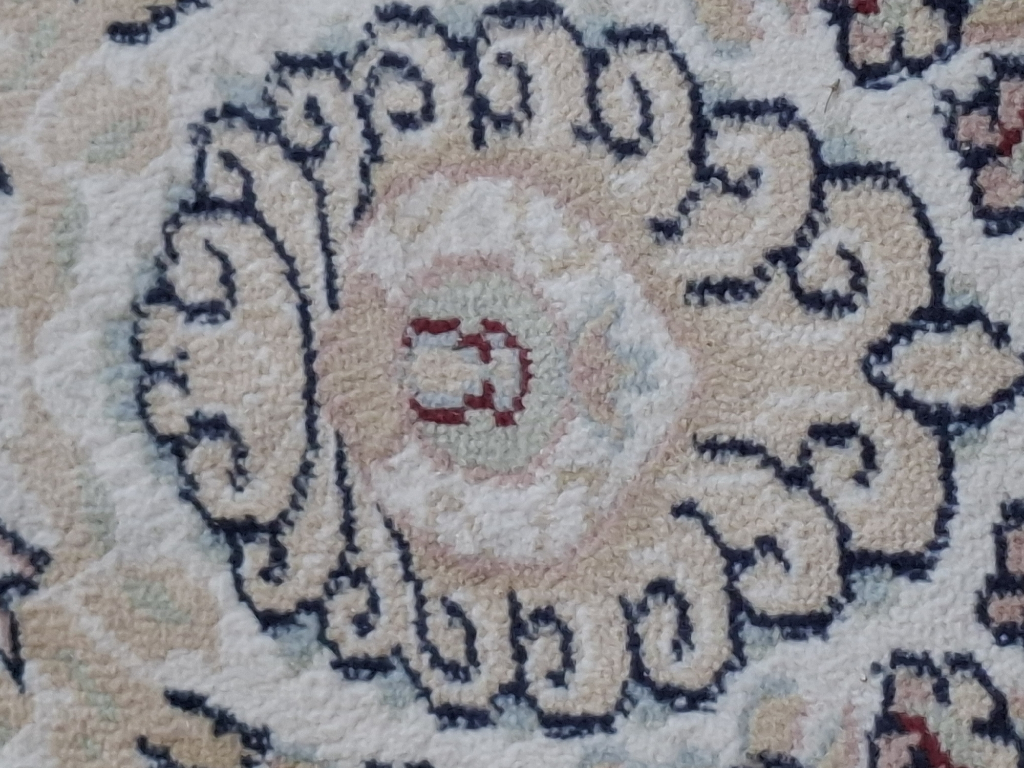The image displays a close-up, overhead view of a rug with a repeating floral or medallion pattern. The pattern features swirls of beige and cream, outlined in dark blue, with touches of muted red and pale green interspersed. The rug's texture appears soft and plush. No information is available to determine the location in Almaty, Kazakhstan, as the image only shows the rug itself without any contextual environmental or architectural cues.
