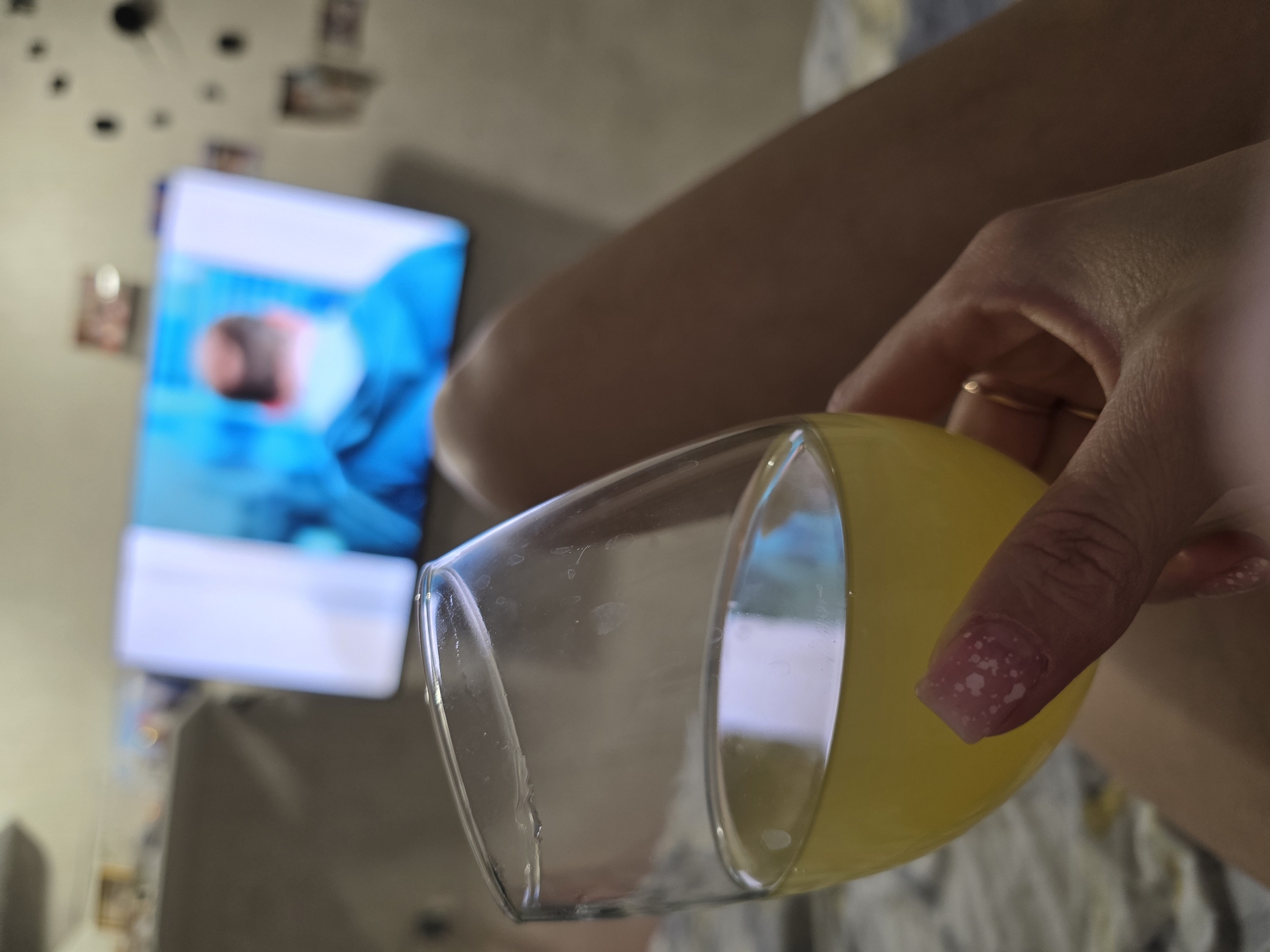 A close-up, slightly tilted shot shows a hand holding a glass filled with yellow liquid, likely juice. The hand has painted nails with a white speckled design. A gold ring is visible on one finger. The background is blurred but shows a television screen displaying an image of a person in blue attire and an out-of-focus wall with decorations. It is impossible to determine from this image if the scene is in the city of Chaikovsky, Russia or any specific location.