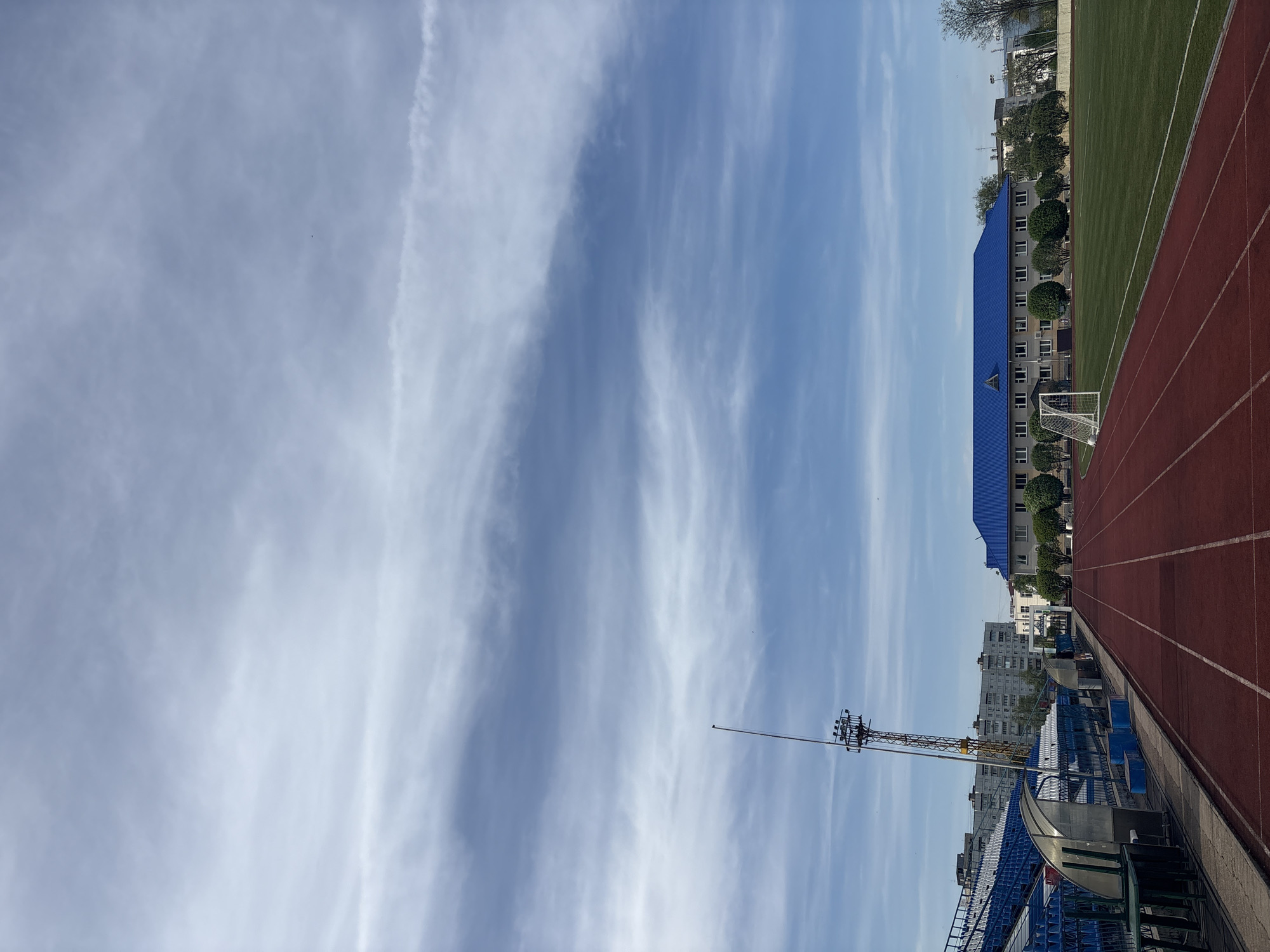 The image depicts an athletic stadium in Chaikovsky, Russia, under a sky filled with wispy clouds. The stadium features a red running track and a green playing field. A blue-roofed building runs alongside the track, partially obscured by trees. In the foreground, there are blue spectator seats. A tall communications tower rises between the seating area and the sky. Buildings in the city are visible in the background behind the stadium.