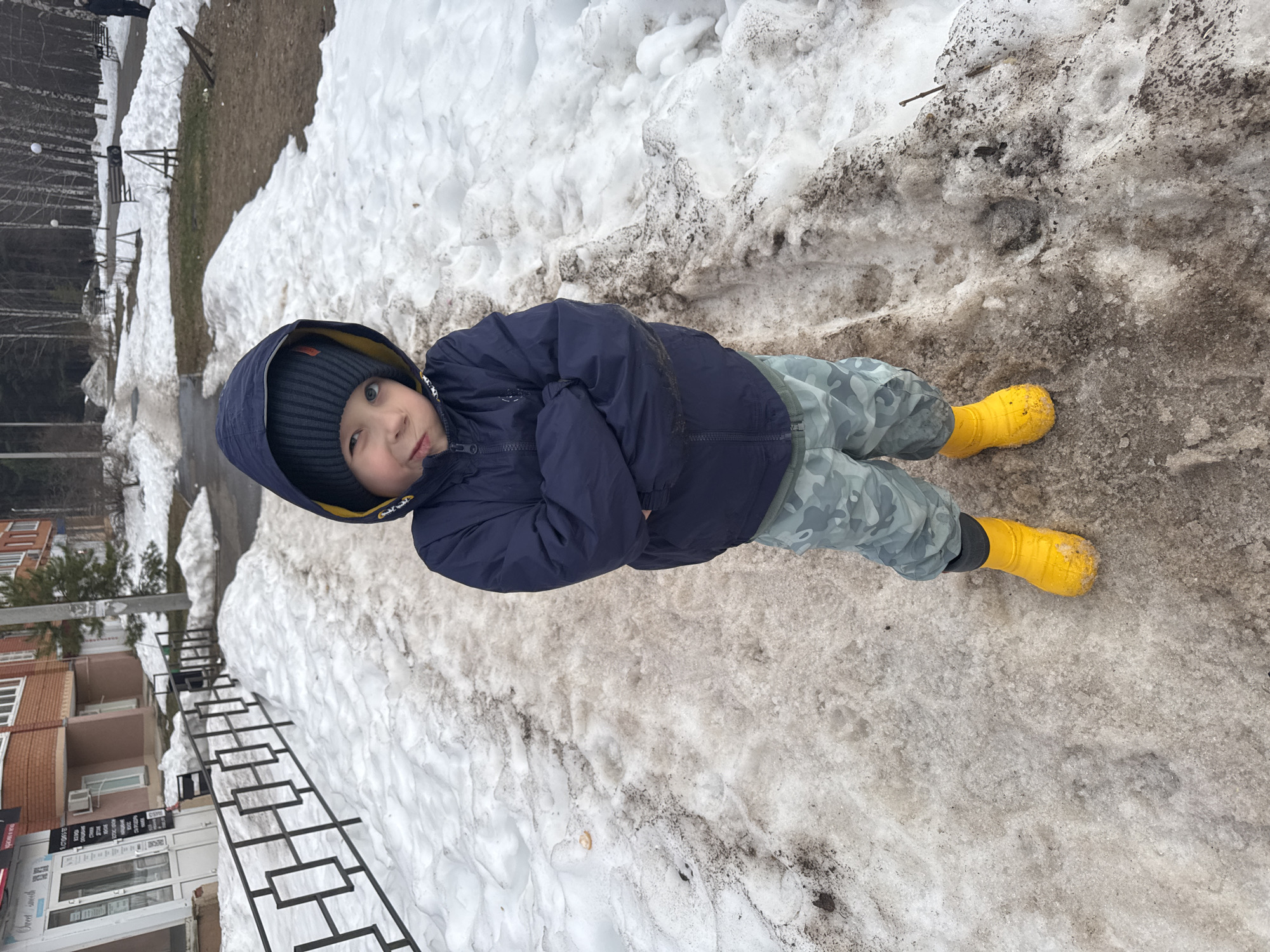 A young child in a blue hooded jacket, camouflage pants, and yellow boots is lying in dirty, partially melted snow. The location appears to be an urban setting in Chaikovsky, Russia, evidenced by a multi-story brick building with a store front in the background and a metal railing running along the sidewalk. The child is positioned with their head slightly elevated. The overall environment suggests late winter or early spring.