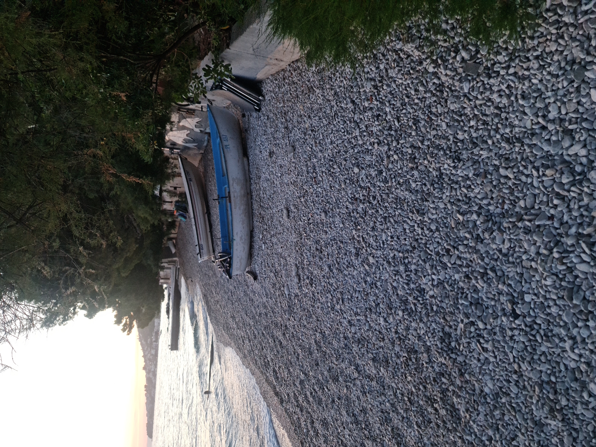 The image shows a pebble beach in Brist, Croatia. Two small boats are pulled up onto the shore, one white with a blue trim and the other white with a registration number "MA 1797". A person is visible swimming in the shallow water near the shore. The sea is calm, with gentle waves lapping against the beach. Trees and vegetation line the edge of the beach, casting shadows on the pebble-covered ground. A concrete retaining wall runs along the back of the beach. The sky is bright with the soft light of either dawn or dusk.