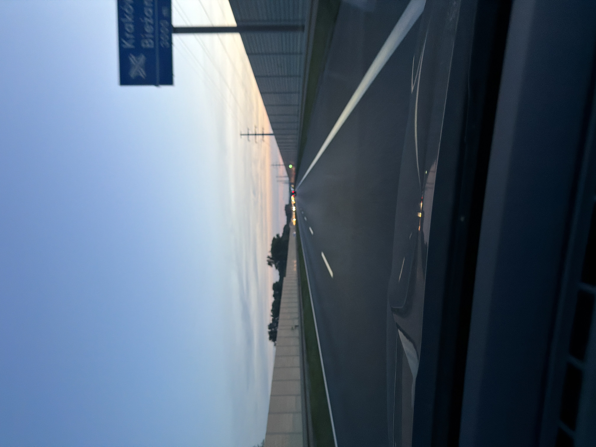 The image shows a view from inside a vehicle driving on a highway at dusk. A blue sign is visible on the right side of the road, indicating "Kraków" and "Biezanów" with distances of 3000 meters. The highway has multiple lanes and is bordered by sound barriers. The sky is clear blue, transitioning to a softer gradient of orange and pink near the horizon. Distant traffic lights and streetlights are visible in the distance. The foreground shows the interior of the vehicle, including the dashboard. The location is identified as Kokotów, Poland, along a highway leading towards Kraków.