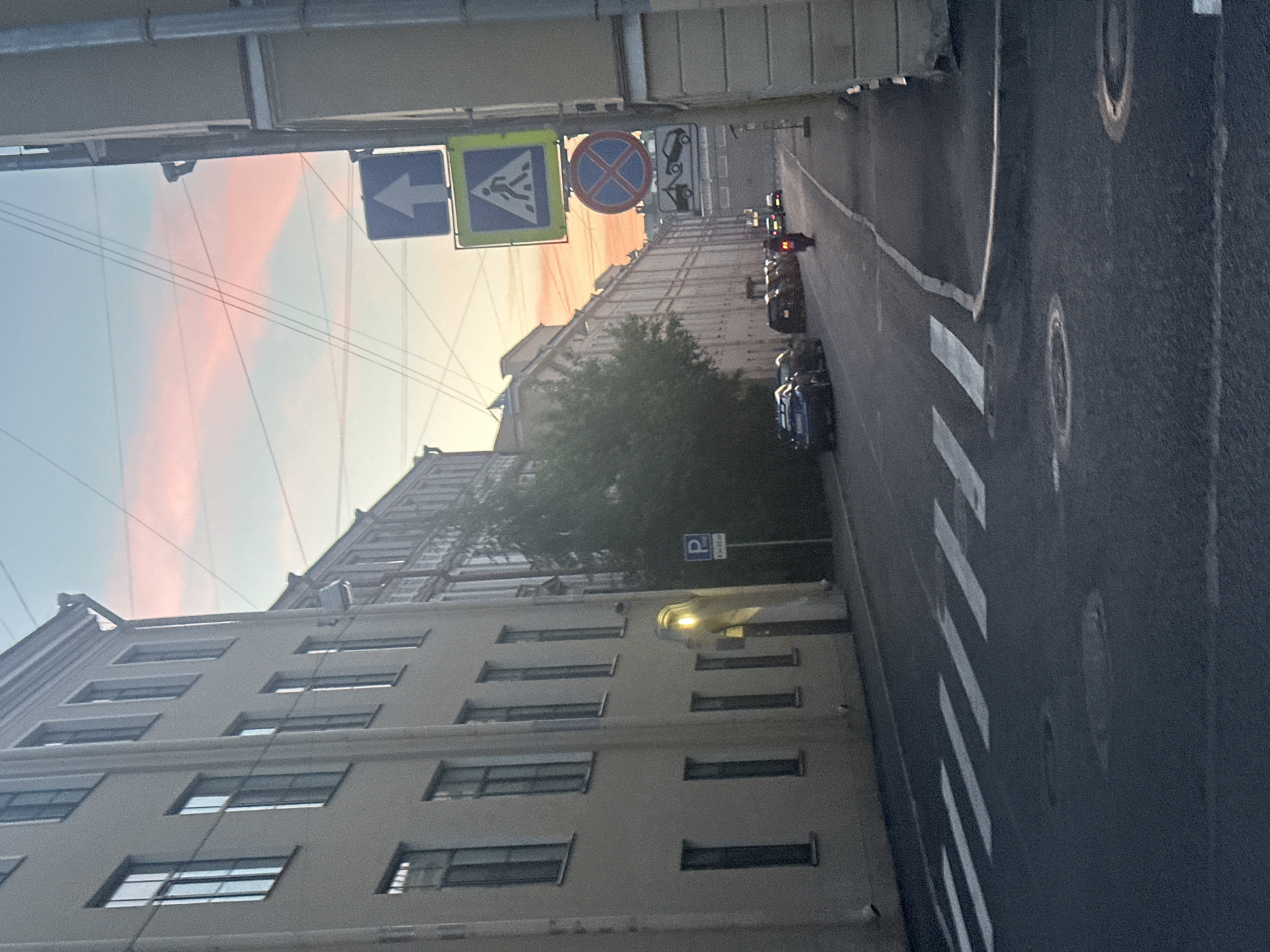 In the city of Unknown, Unknown, a street scene unfolds under a sky painted with soft pink and orange hues of sunset. Tall, cream-colored buildings with numerous windows line the left side of the asphalt road. Across the street, another building stretches into the distance. A marked pedestrian crossing is visible on the road. Several cars are parked along the side of the street. A motorcycle rider is proceeding down the street. Traffic signs, including a pedestrian crossing sign, a "no stopping" sign, and a towing sign, are posted on the right side of the road. Power lines crisscross the sky.