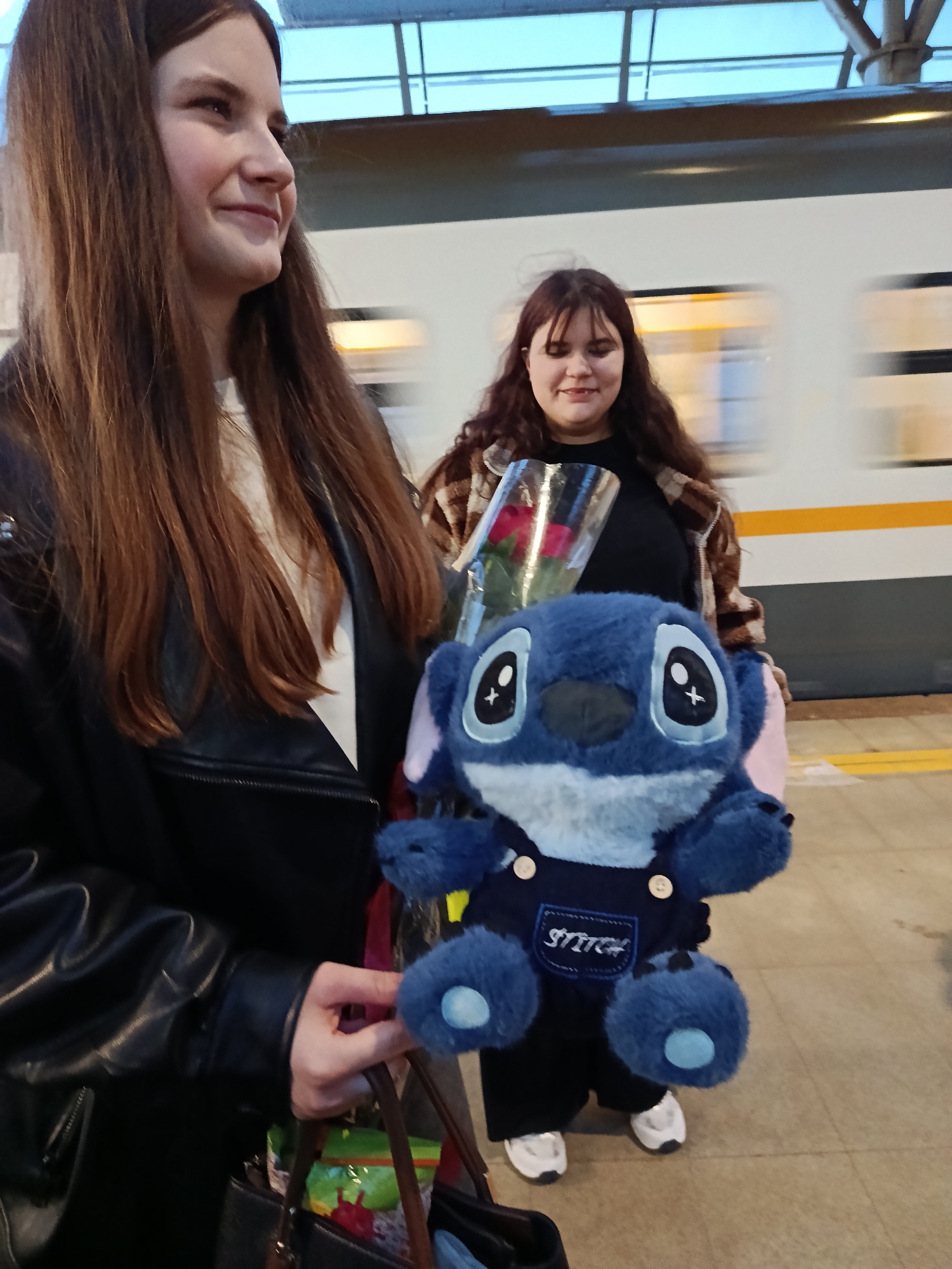 Two women stand on a train platform. The woman in the foreground, wearing a black leather jacket, holds a Stitch plush toy and a brown purse. Behind her, another woman with brown hair is wearing a brown plaid coat, holding a bouquet of red roses. A train passes in the background. The location is likely a train station, possibly in Moscow, Russia, given the architectural style of the overhead structure.