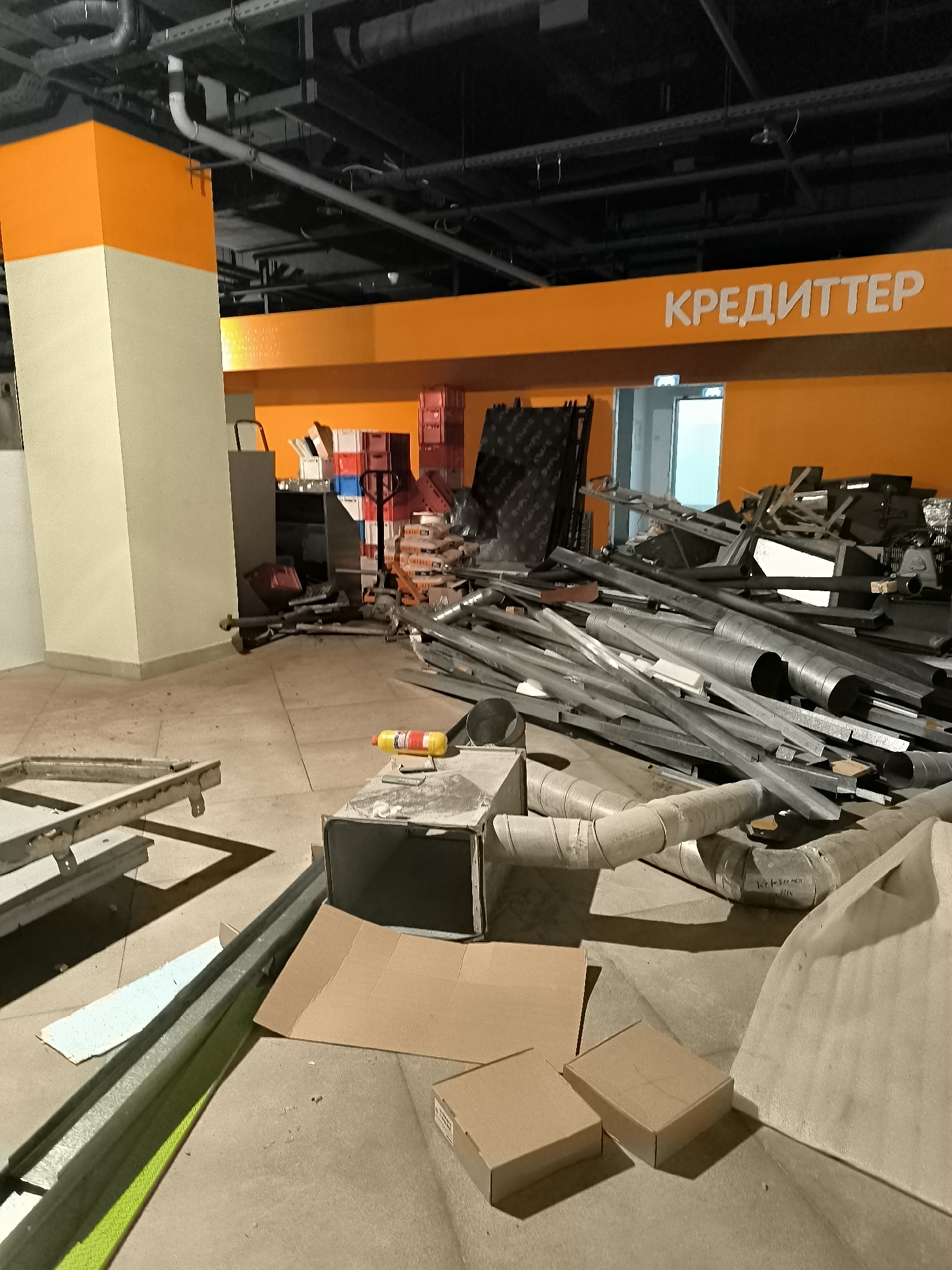 An interior view shows a construction or renovation area within a building, possibly a retail space, given the prominent orange signage with Cyrillic text that reads "КРЕДИТТЕР" (CREDITTER). The floor is concrete, and various construction materials are piled haphazardly. Dominating the scene are large quantities of metal ducting, pipes, and structural elements, suggesting an HVAC or infrastructure project. Cardboard boxes and other debris are scattered around. In the background, an orange and light beige pillar stands, and more stacked boxes and equipment are visible. The lighting is artificial and somewhat dim. There are no people present in the image. The location is not specified in the image.