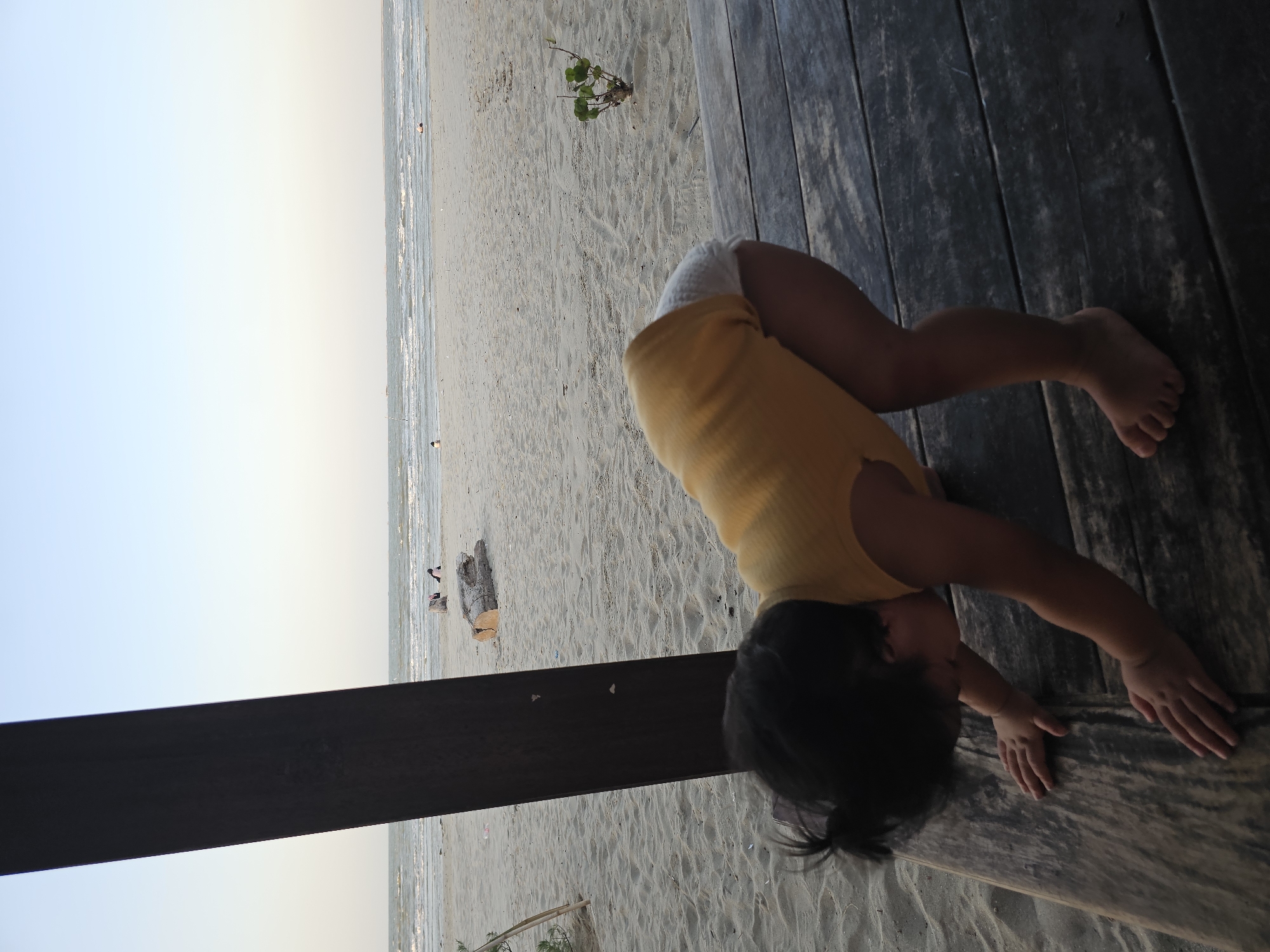 The media file shows a young child in a yellow shirt and white diaper attempting a yoga pose on a wooden deck. The child is positioned upside down with their hands on the deck and their legs in the air. The setting is a sandy beach with the ocean in the background. The sky is a pale blue, suggesting it is either early morning or late afternoon. There are a few scattered pieces of driftwood on the beach, and some footprints in the sand. The overall mood is playful and relaxed. The image is taken from a high angle, looking down at the child. There is no visible text in the image.