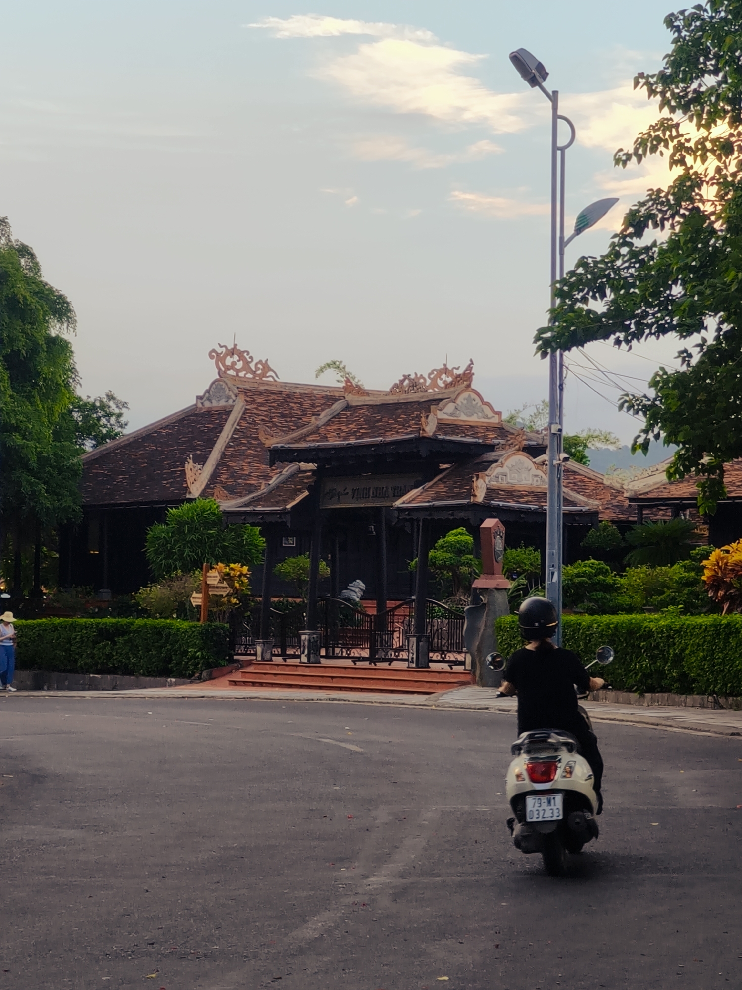 A person rides a white scooter down a paved road in front of a traditional Vietnamese building. The building has an ornate, tiled roof with intricate carvings and is surrounded by lush green bushes and trees. A street lamp stands to the right of the building, and a person wearing a straw hat is visible on the left, standing near the bushes. The sky is a hazy blue with scattered clouds, suggesting it is either early morning or late afternoon. The scene appears peaceful and scenic, with the architectural style of the building indicating a cultural or historical location in Vietnam. The license plate on the scooter reads "79-M1 032.33".