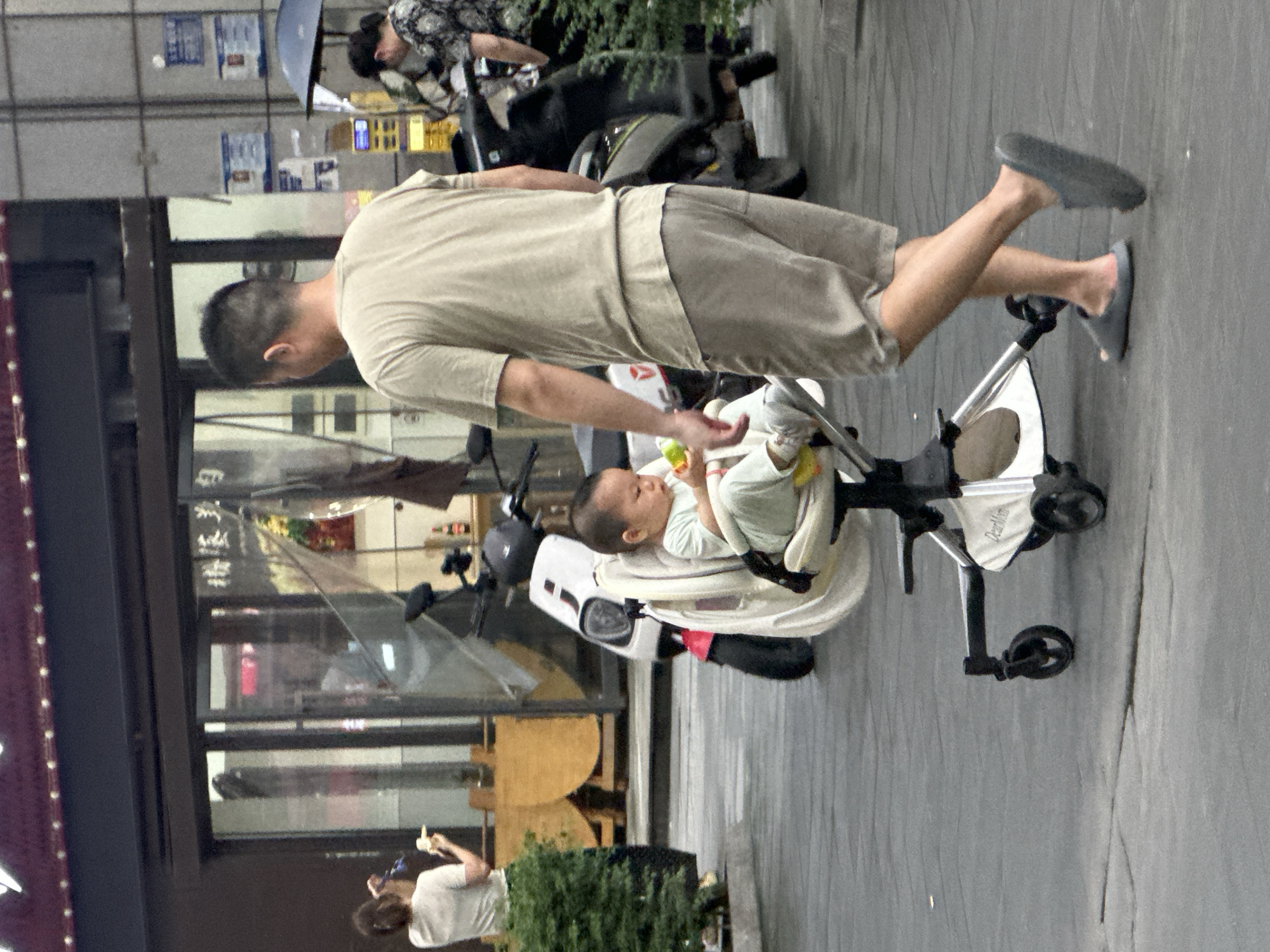 This image shows a street scene in Chongqing High-tech Zone, China. The primary subjects are a man, a baby, and a baby stroller. The man is pushing the stroller, which contains a baby. The scene appears to be outdoors, likely on a sidewalk, with a building in the background. There are also elements of urban street life visible, including what looks like a motorcycle and possibly storefronts.
The activity taking place is a man pushing a baby in a stroller. It appears to be a casual outing. The time of day is not definitively clear, but the lighting suggests it could be daytime, possibly overcast. No specific weather conditions are apparent, though the ground looks a bit wet, which could indicate recent rain.
Notable details include the man's attire - a light-colored t-shirt and shorts, and the baby's light-colored clothing. The stroller is white and black with silver accents. There is some text visible on the stroller, which appears to say "DearMom". Other signs and writing are visible in the background, but are too blurry to decipher. The overall mood is calm and everyday.