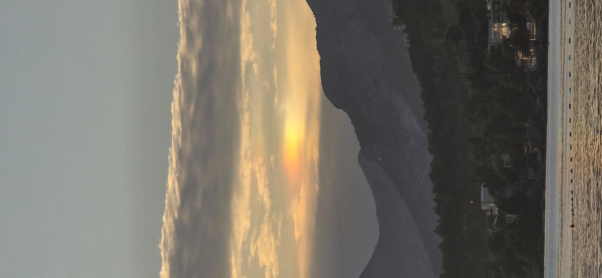 The image depicts a coastal scene during either sunrise or sunset over the city of Bar, Montenegro. The sky features a light blue-grey upper section transitioning to warm golden tones below, with horizontal cloud bands. A prominent iridescent optical phenomenon, displaying red, orange, yellow, and green hues, is visible within the golden cloud layer towards the upper-left of the obscured sun. Multiple layers of dark, silhouetted mountains occupy the mid-ground, receding into the distance. The foreground is dominated by a calm body of water, reflecting the golden sky light in shimmering patterns. A line of approximately ten dark cylindrical buoys and one red spherical buoy are present on the water's surface, extending from the mid-left towards the right. On the lower slopes and coastline, several buildings are observed amidst dark foliage, with some interior lights visible through windows. No human figures or discernible actions are present.