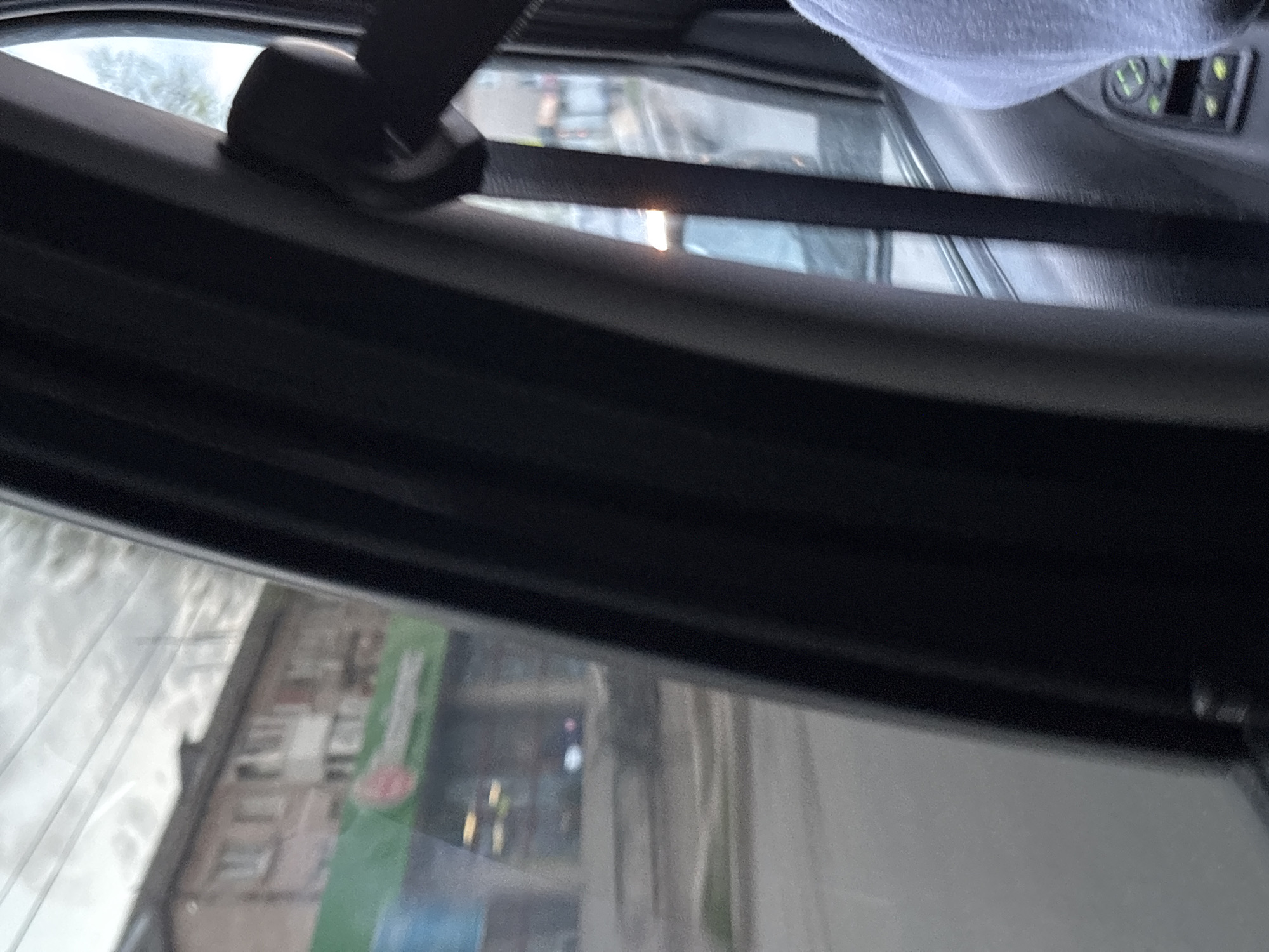 A view from inside a vehicle, looking out through a window. The scene reflects a city street with buildings visible in the background, including a building with green signage and red accents. This photo was taken in Chaikovsky, Russia. Parts of the car's interior are visible, including the black trim, a seatbelt mechanism, and what appears to be a light-colored cloth or headliner at the top of the frame. The angle suggests the photo was taken from a low vantage point within the vehicle. The focus is on the exterior view, although it is slightly blurred due to the glass and motion.