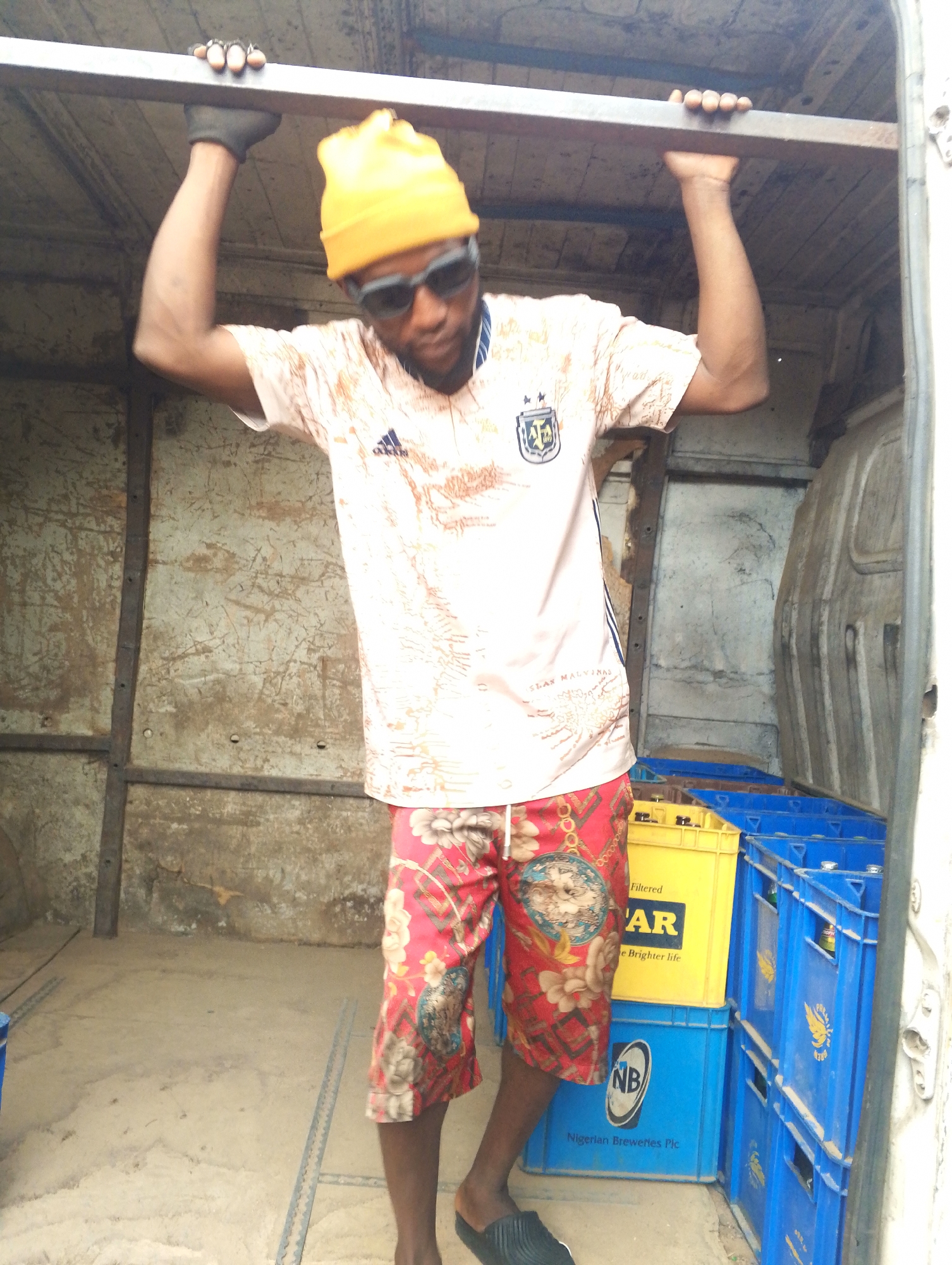The image shows a man with dark skin inside a vehicle, possibly a van or truck. He is wearing a yellow beanie, sunglasses, a light pink T-shirt with an Argentinian football team emblem, and red floral shorts. His hands are gripping a metal bar above his head. He is wearing black gloves on his hands. Behind him, there are stacks of blue and yellow crates, some bearing the branding of "Star" beer and "Nigerian Breweries Plc." The interior of the vehicle appears worn. The location is unknown.