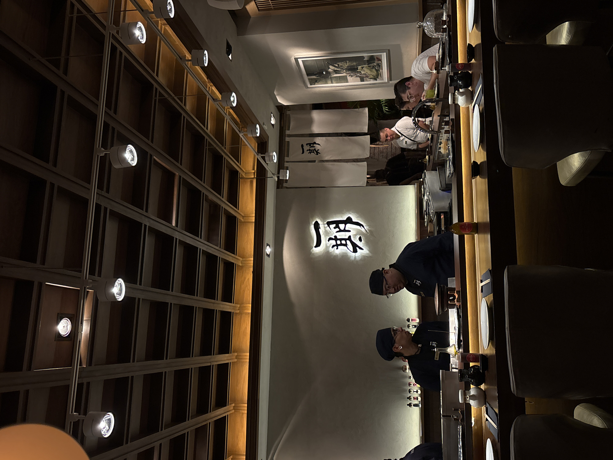 This media file captures the interior of a sophisticated Japanese restaurant, likely a sushi bar, during the evening. The scene is dimly lit, creating an intimate ambiance.
At the center of the image, a long wooden counter runs horizontally, serving as the main focus. On the right side of the counter, two chefs, dressed in dark blue uniforms and caps, are visible. They appear focused on their work or interacting with guests, leaning slightly forward. On the opposite side of the counter, on the left, several customers are seated. One man in a white shirt is attentively looking down at the counter, while another is holding a glass to his mouth, possibly drinking.
The background wall, behind the customers and chefs, features a large, glowing sign with prominent Japanese characters that appear to read "一麗" (Ichirei or similar). Above the counter, three white banners are hung, each displaying Japanese characters; one clearly shows the character "一". Further left, a framed piece of artwork hangs on the wall.
To the far left of the image, a tall wall is adorned with a grid-like wooden structure, possibly shelving or decorative paneling, illuminated by multiple track lights. Similar track lighting fixtures are also visible on the ceiling, casting a warm glow throughout the space. The counter itself is set with plates, chopsticks, and small serving dishes, along with various bottles (possibly sake or condiments) and other barware. The overall decor is modern and minimalist, with a strong emphasis on wood textures.
The setting suggests an upscale dining experience in a Japanese restaurant, located in Mexico City, Mexico.