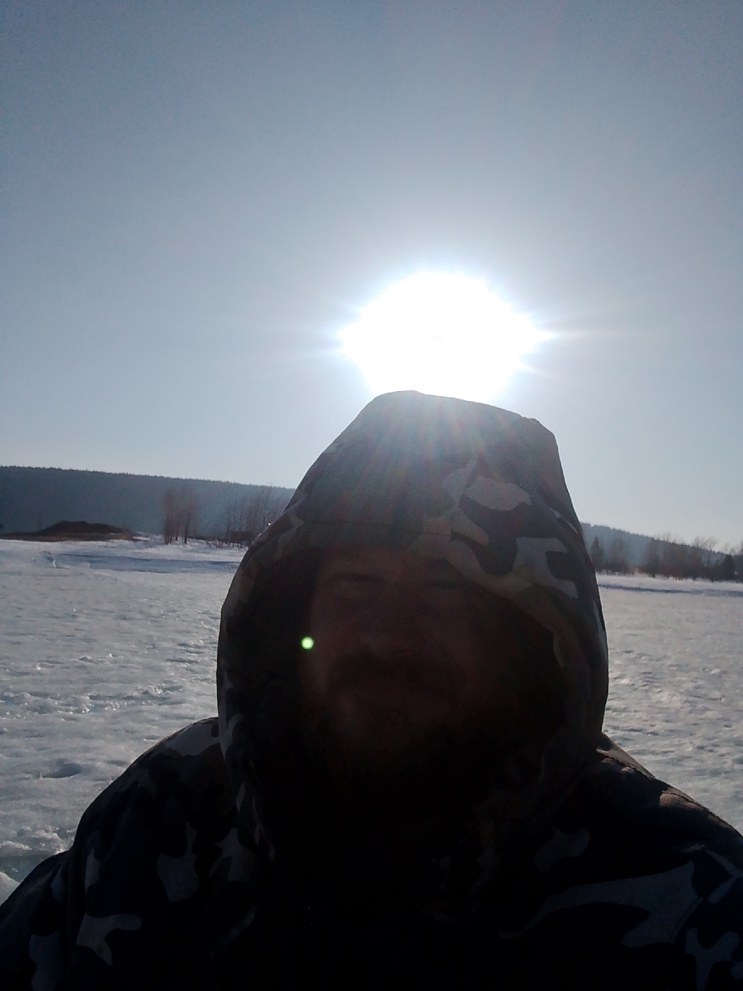 A person wearing a camouflage-patterned hooded jacket stands in a snowy environment under a bright sun. The landscape appears to be a snow-covered field or frozen body of water with sparse vegetation in the distance. The location is Noviy, Russia.