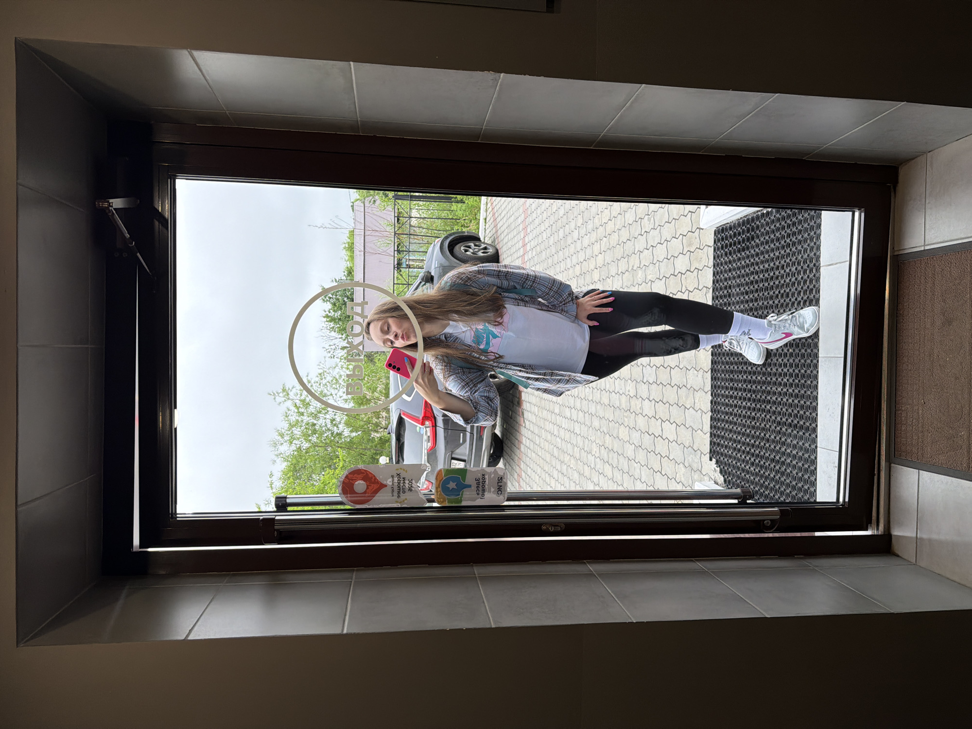 A young woman is captured in a mirror selfie, standing in a doorway. She is wearing a light-colored t-shirt with a graphic print, a plaid shirt, black leggings, white socks, and grey and pink athletic shoes. Her hair is long and brown, and she is holding a pink smartphone in her right hand. Her lips are pursed. The doorway appears to be an entrance to a building, possibly a shop or restaurant, with a sign visible in the reflection that reads "ВЫХОД" (EXIT) in Cyrillic letters. Two stickers are adhered to the glass door, one with a red pin marker and another with a blue star. Outside the door, a paved area is visible, with a parked dark-colored SUV and some greenery. The scene is likely set in Birobidzhan, Russia, given the Cyrillic text on the sign. The lighting suggests it is daytime, with an overcast sky.