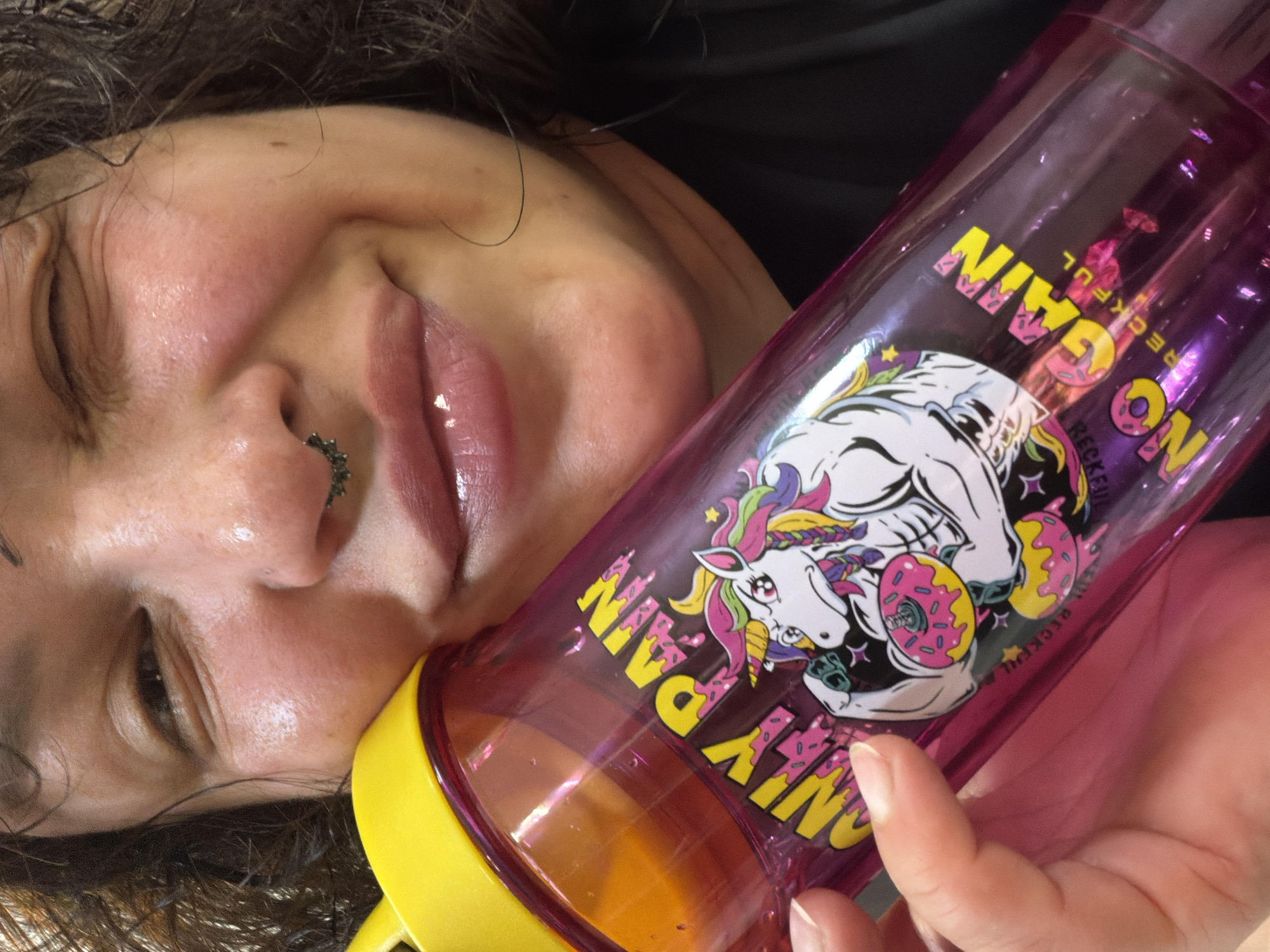 The image shows a close-up of a person's face and a pink, translucent water bottle with a yellow lid. The person is holding the bottle up to their cheek, with their left eye partially closed and a slight smile. The water bottle has a graphic design that includes a muscular unicorn holding two donuts, and the text "ONLY PAIN NO GAIN" and "RECKFUL". The background is dark. The image does not contain any information about the location in Chaikovsky, Russia.