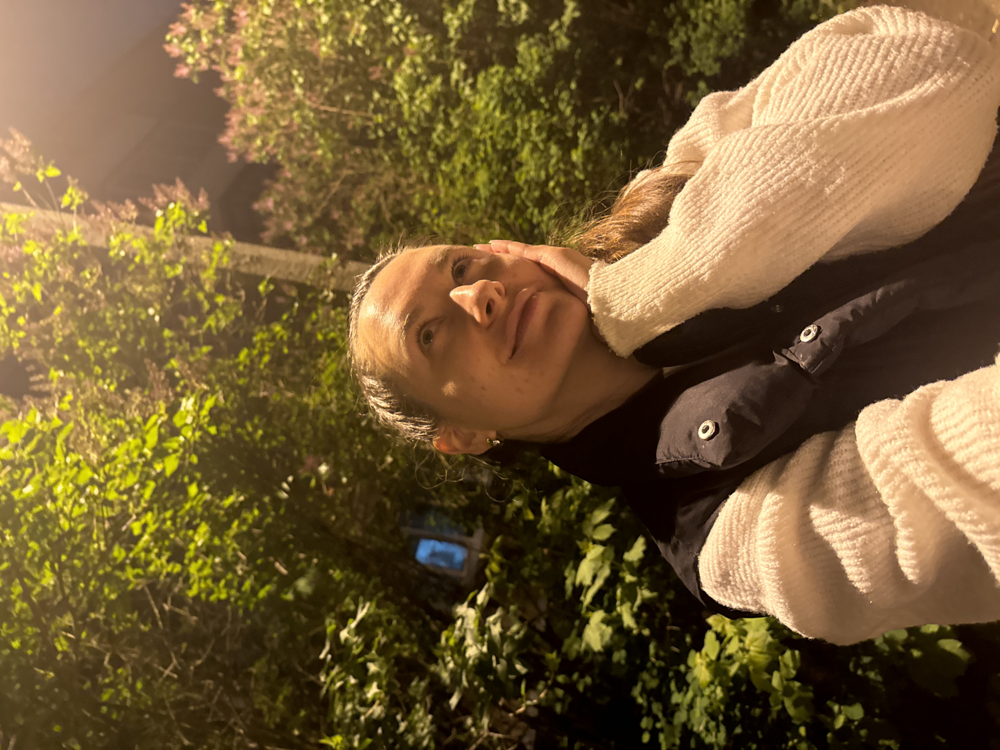 A woman is standing outside at night in Vniissok, Russia. She is wearing a white sweater over a black coat. The background is filled with lush green foliage and glimpses of buildings. She has her hand to her face. The lighting is dark, illuminated by artificial light sources.
