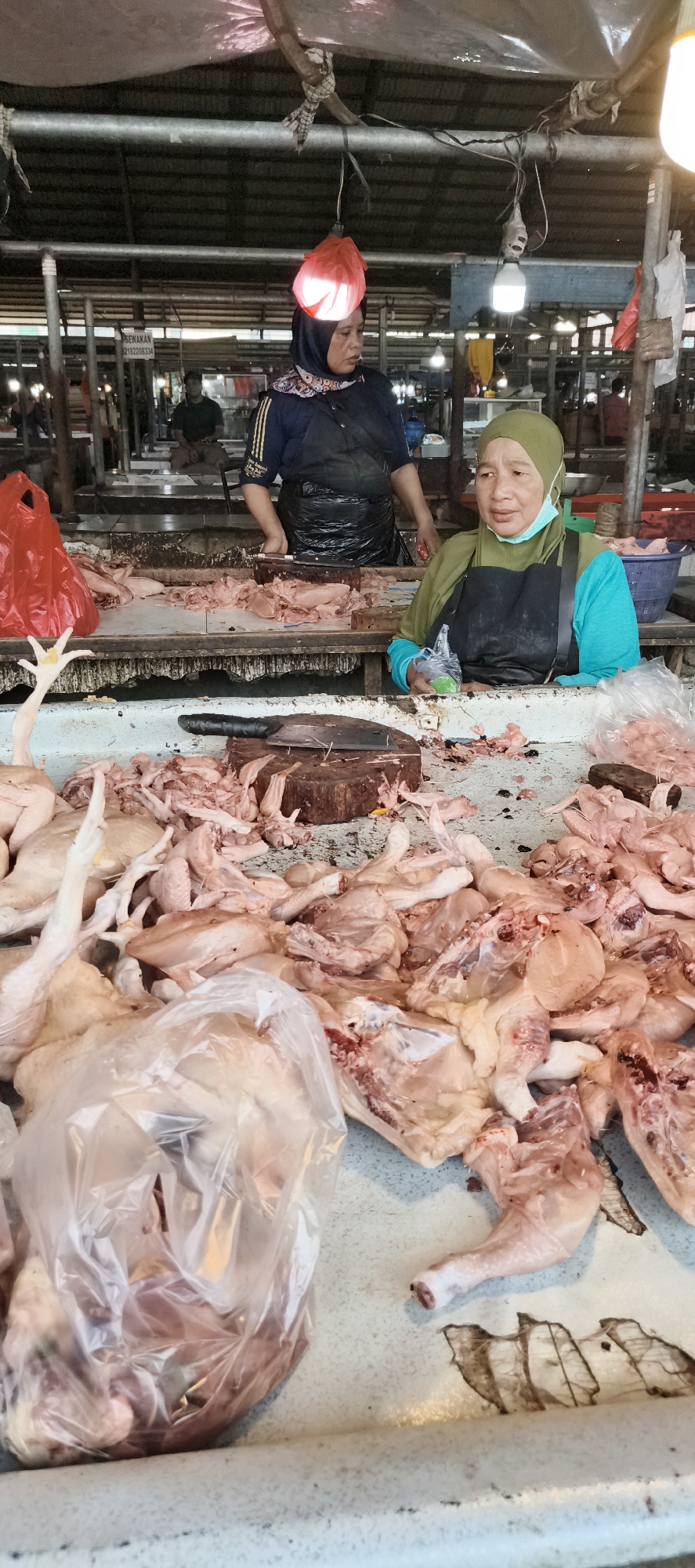 The image depicts a busy marketplace scene in Jambi, Indonesia, likely a traditional market with an outdoor or semi-outdoor setting. Two women are the primary subjects, both appearing to be vendors.
The woman on the left, wearing a dark blue top, black apron, and a patterned headscarf, is partially obscured by the stacked chicken pieces. She is standing behind a counter and appears to be actively working, possibly preparing or displaying the poultry.
The woman on the right, who is more prominently featured, wears a bright green headscarf and a turquoise shirt, with a black apron over it. She is looking towards the viewer or slightly to the right, with a neutral expression. She is also positioned behind a counter laden with a large quantity of raw chicken parts, including whole chickens, legs, and other pieces, some of which are wrapped in plastic bags. A light blue disposable face mask is visible around her neck.
The foreground is dominated by the raw chicken meat, which is displayed on a white surface that appears to be a counter or table. There are also various tools visible, including a large cleaver on a wooden block, suggesting the processing of the poultry is taking place. Red plastic bags are also present, one of which is prominent on the left side of the image.
The background reveals more of the market environment. There are structures that look like stalls or market stalls, with tarpaulin-like coverings. Other people are visible in the background, some appearing to be customers or other vendors, though they are less distinct. Lighting in the market appears to be a mix of natural light and artificial sources, such as hanging bulbs. The overall impression is one of a vibrant, active food market.