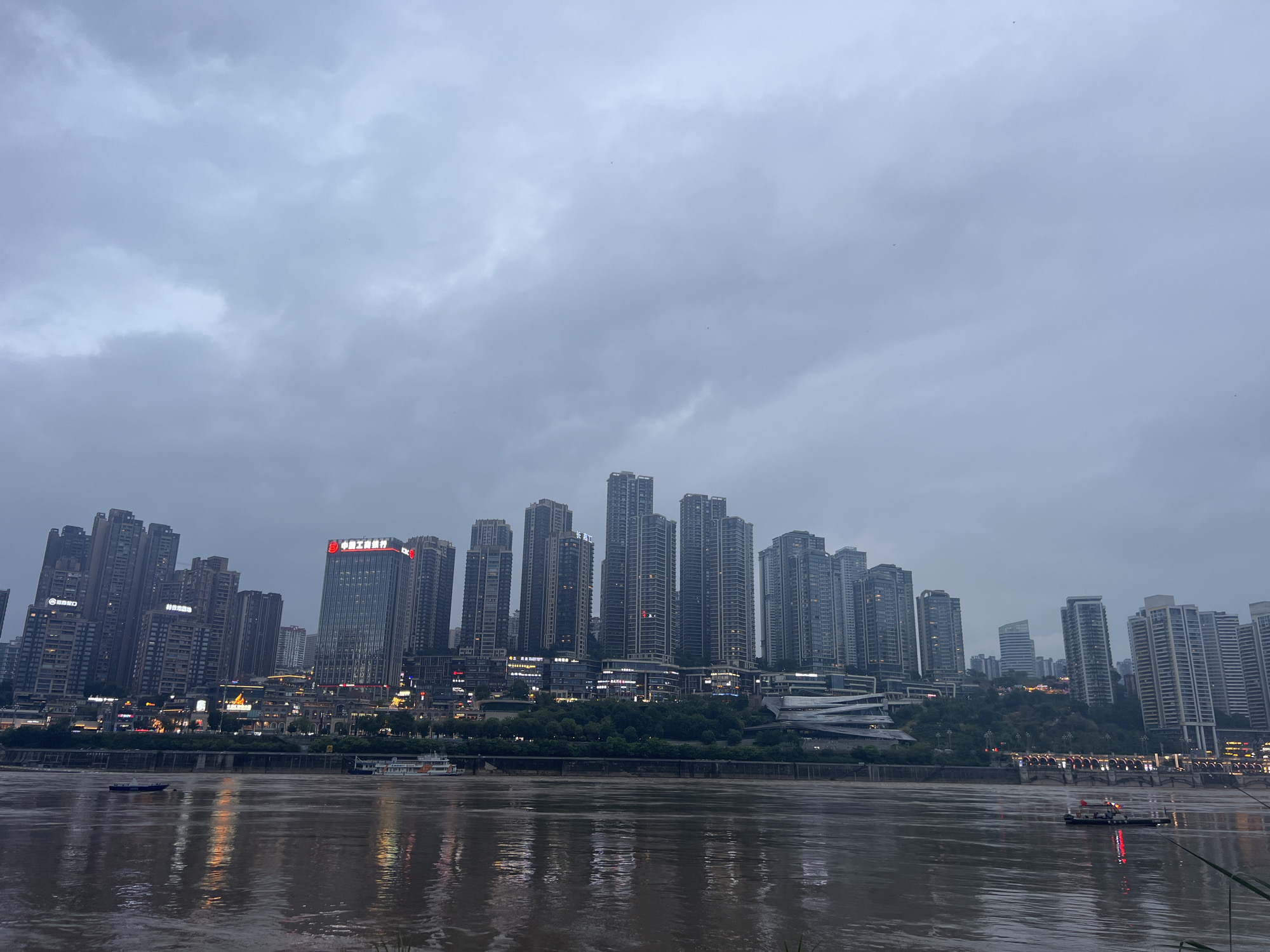 The image depicts a waterfront cityscape at dusk, likely in Nan'an District, China, given the distinct architecture and urban density. A wide, murky river dominates the foreground, reflecting the lights of the city buildings. Two small boats are visible on the river. The middle ground features a densely packed skyline of numerous high-rise buildings, with some illuminated windows indicating internal activity. A prominent red sign with Chinese characters is visible on one of the taller buildings. A tree-lined embankment separates the river from the urban development. The sky is overcast and gray, suggesting twilight or impending rain. The overall impression is one of a bustling, modern Chinese city.