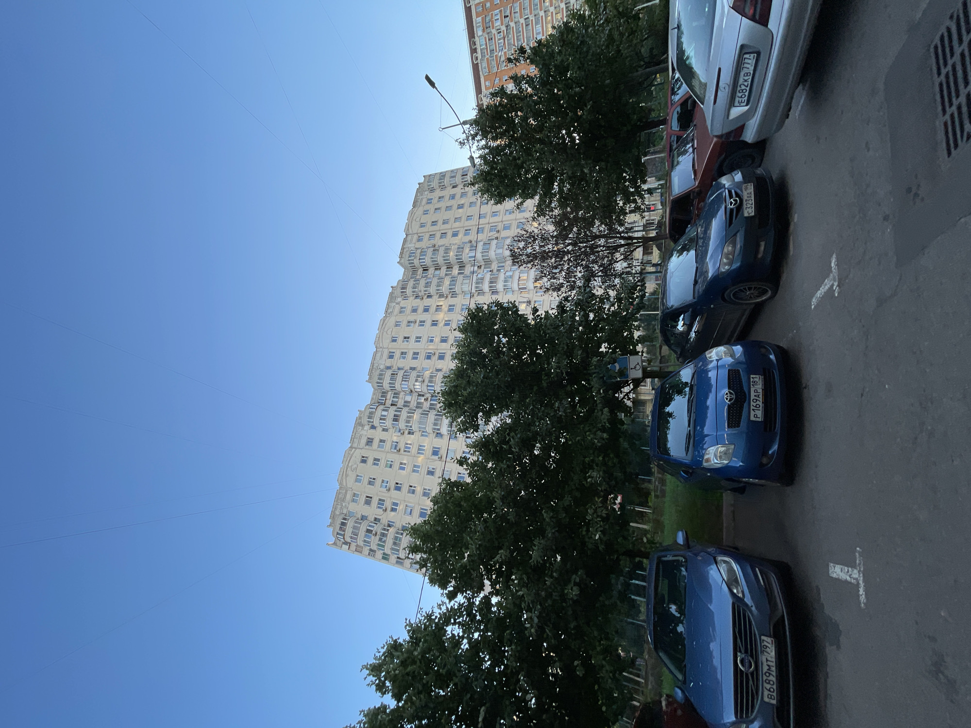 This media file depicts a street scene in Moscow, Russia, with a clear blue sky overhead. The primary subjects are several parked cars and a tall, light-colored residential building with a distinctive curved facade, featuring numerous balconies and windows. Trees with lush green foliage are interspersed between the building and the street, casting some shade.
The cars are parked on a paved surface, and parking lines are visible on the ground. The time of day appears to be late afternoon or early evening, given the angle of the sunlight and the shadows. The weather seems pleasant, with a cloudless sky.
Visible text includes license plates on the cars. One license plate reads "E682KB 777", another "C323AA 99", and a third "P169AP 181", and the last one "B689MT 797". The overall atmosphere is one of everyday urban life.