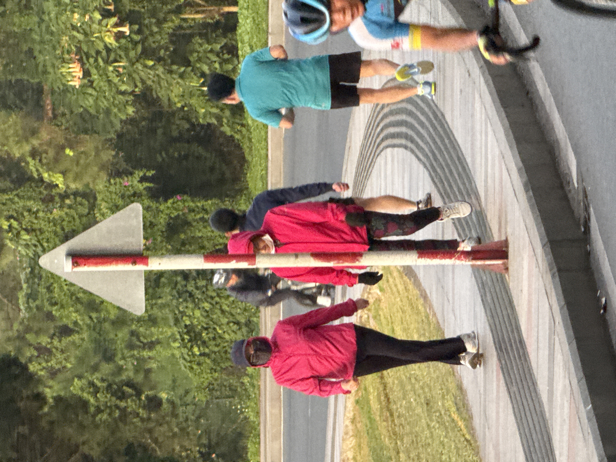 The image captures a pedestrian walkway adjacent to a road in Da Lat, Vietnam. A triangular sign with red and white stripes stands prominently on the walkway. Several individuals are present: two women in pink hooded jackets, one wearing a black mask and cap, are walking on the path. Further along, two men are jogging, one in a dark long-sleeved shirt and the other in a turquoise t-shirt and black shorts. In the foreground, a cyclist wearing a helmet and blue and yellow cycling gear is visible. The environment is characterized by lush green trees and vegetation bordering the walkway and road. The walkway itself has a distinct textured pattern.