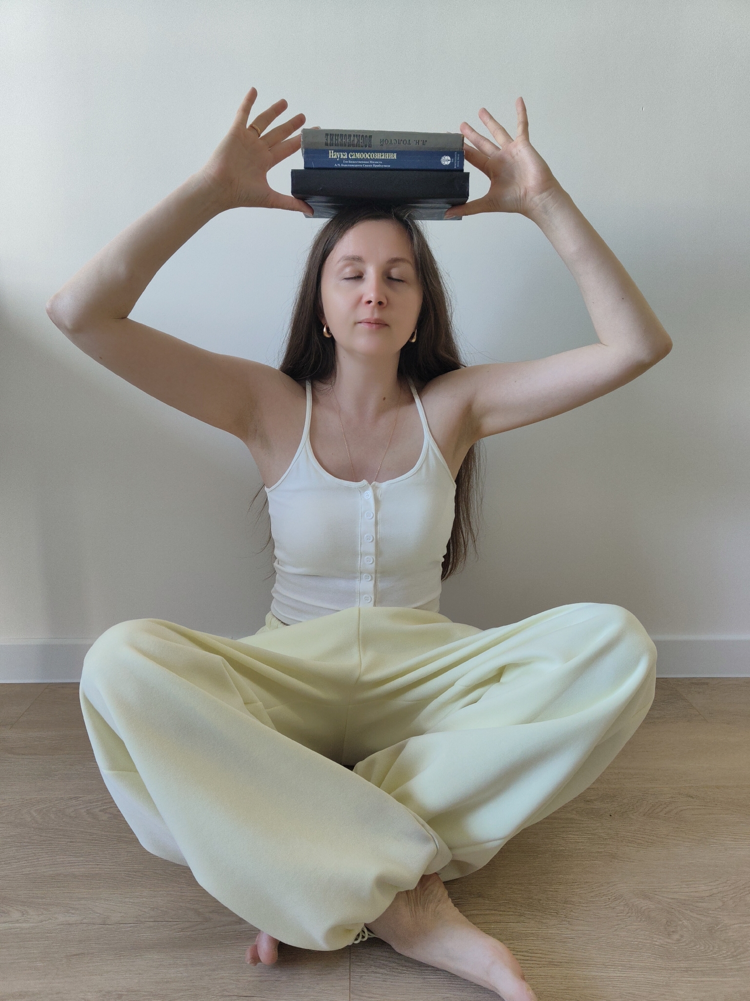 A woman is seated cross-legged on a wooden floor against a white wall in an indoor environment. She is wearing a white tank top and light yellow pants. Her eyes are closed, and she is holding a stack of three books on top of her head with both hands. The books are of varying thickness and color, with some visible text in a Cyrillic alphabet. She is facing forward, with her arms extended above her head. The location is in the city of Unknown, Unknown.