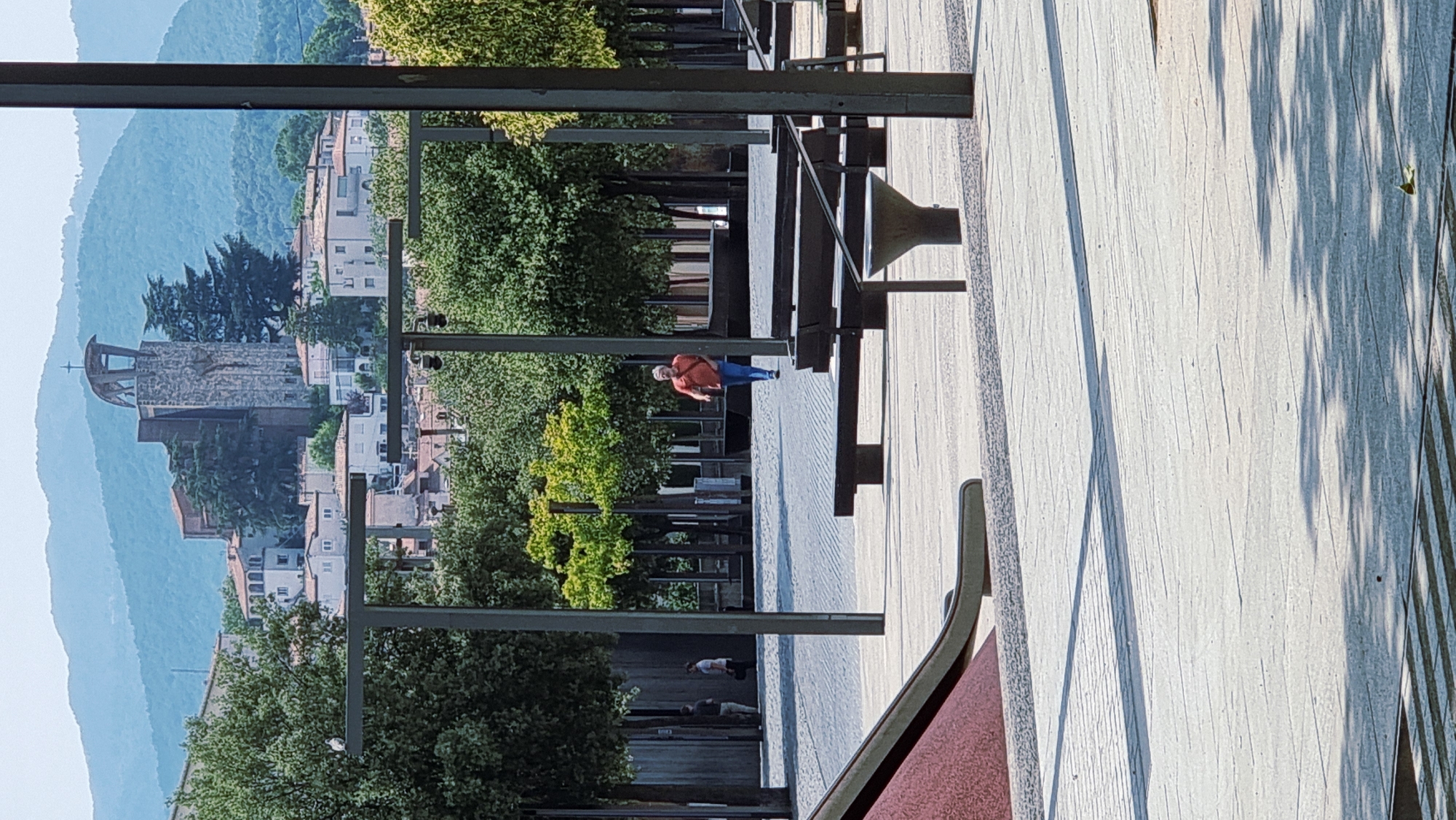 The image depicts an outdoor scene in a city, likely Olot, Spain, characterized by a public plaza with paved ground, trees, and benches. In the background, a distinctive church with a modern bell tower featuring a cross at its apex is visible, situated on a hill overlooking a cluster of buildings. A single man, wearing an orange shirt and blue jeans, is captured mid-stride, walking across the plaza from right to left. The foreground shows a section of a playground, including a red slide. The overall atmosphere is one of a bright, sunny day, with shadows cast by trees and structures on the pavement.
