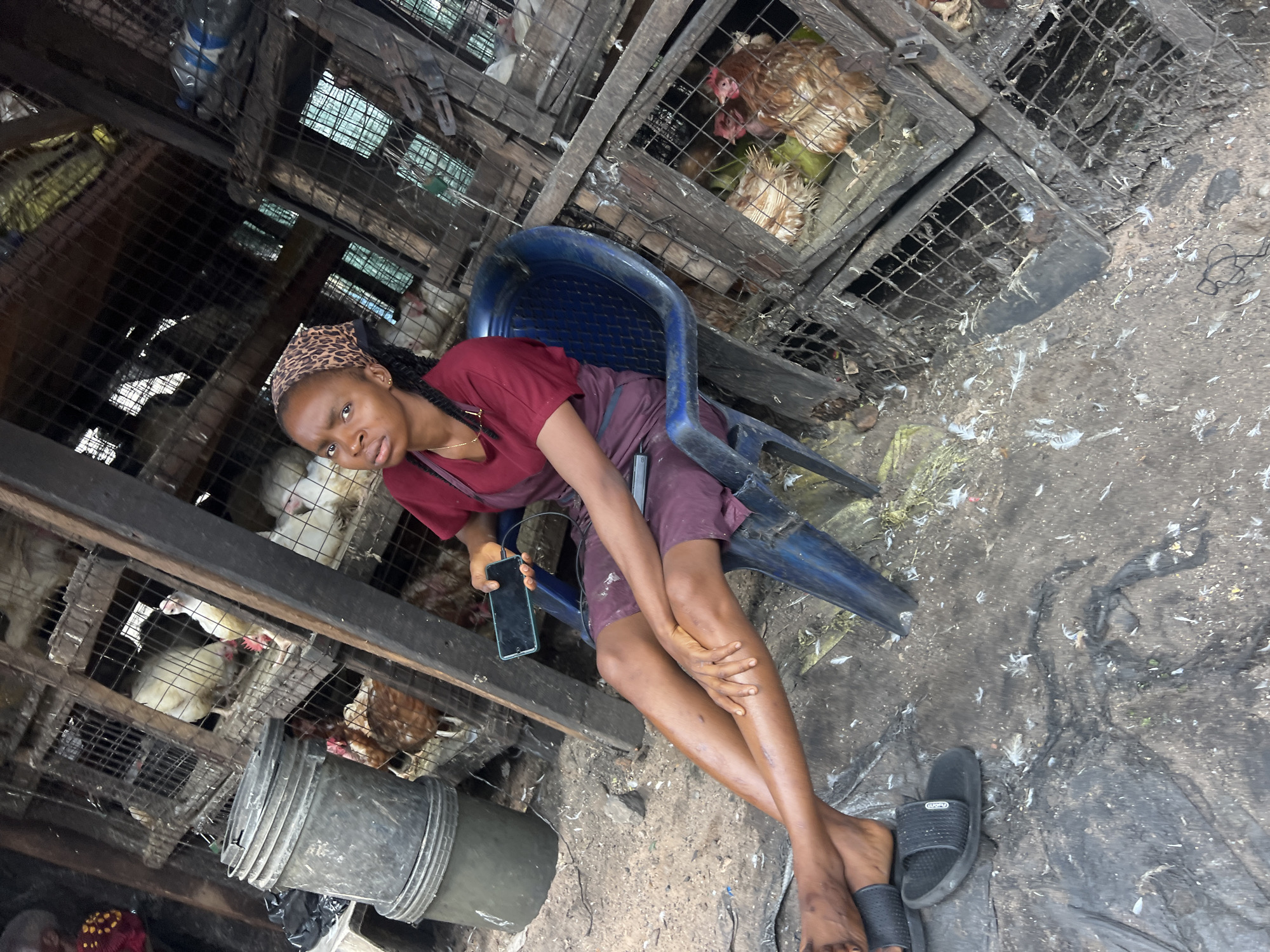 The image depicts a young woman sitting on a blue plastic chair in an outdoor market in Nkpor, Nigeria. She is wearing a maroon top and burgundy shorts, with a leopard-print headscarf. She is holding a smartphone in her right hand. The environment appears to be a poultry market, with numerous wooden cages filled with chickens stacked behind her. The ground is covered with dirt, feathers, and debris. To her left is a large, gray plastic bucket. The overall scene is rustic and indicative of a busy marketplace.