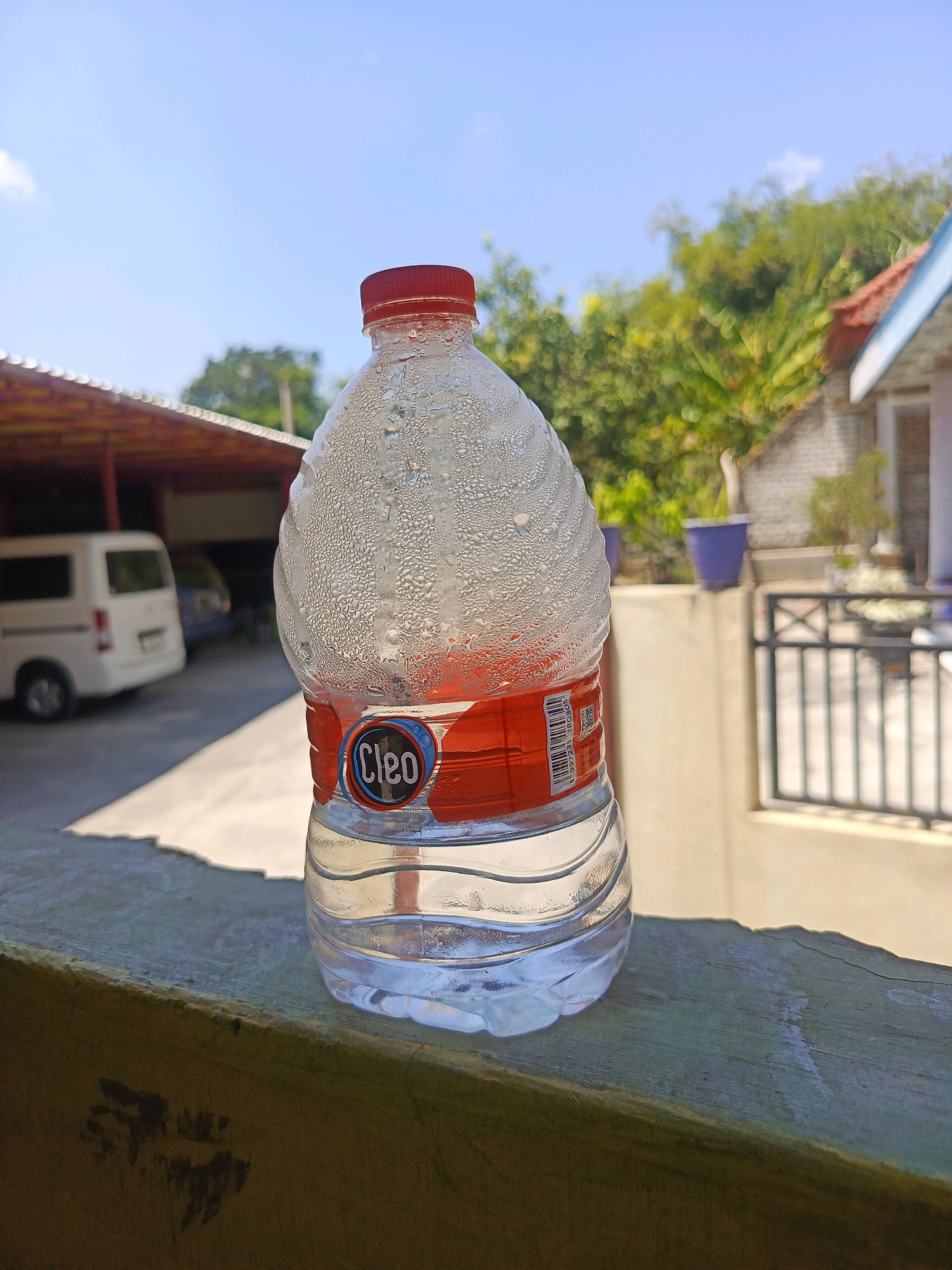 A plastic bottle of water with a red cap sits on a weathered wooden surface. Condensation droplets cover the upper portion of the bottle. The label on the bottle features the brand name "Cleo" in a blue circular emblem against a red background. In the background, a white van is parked under a covered structure with a corrugated roof. Beyond that, trees and a portion of a building with a tiled roof are visible. The scene appears to be outdoors on a sunny day, with a clear blue sky above. The location is likely a residential or commercial area in Pamekasan, Indonesia, given the presence of common Indonesian building styles and a white van, which is a popular vehicle type in the region.
