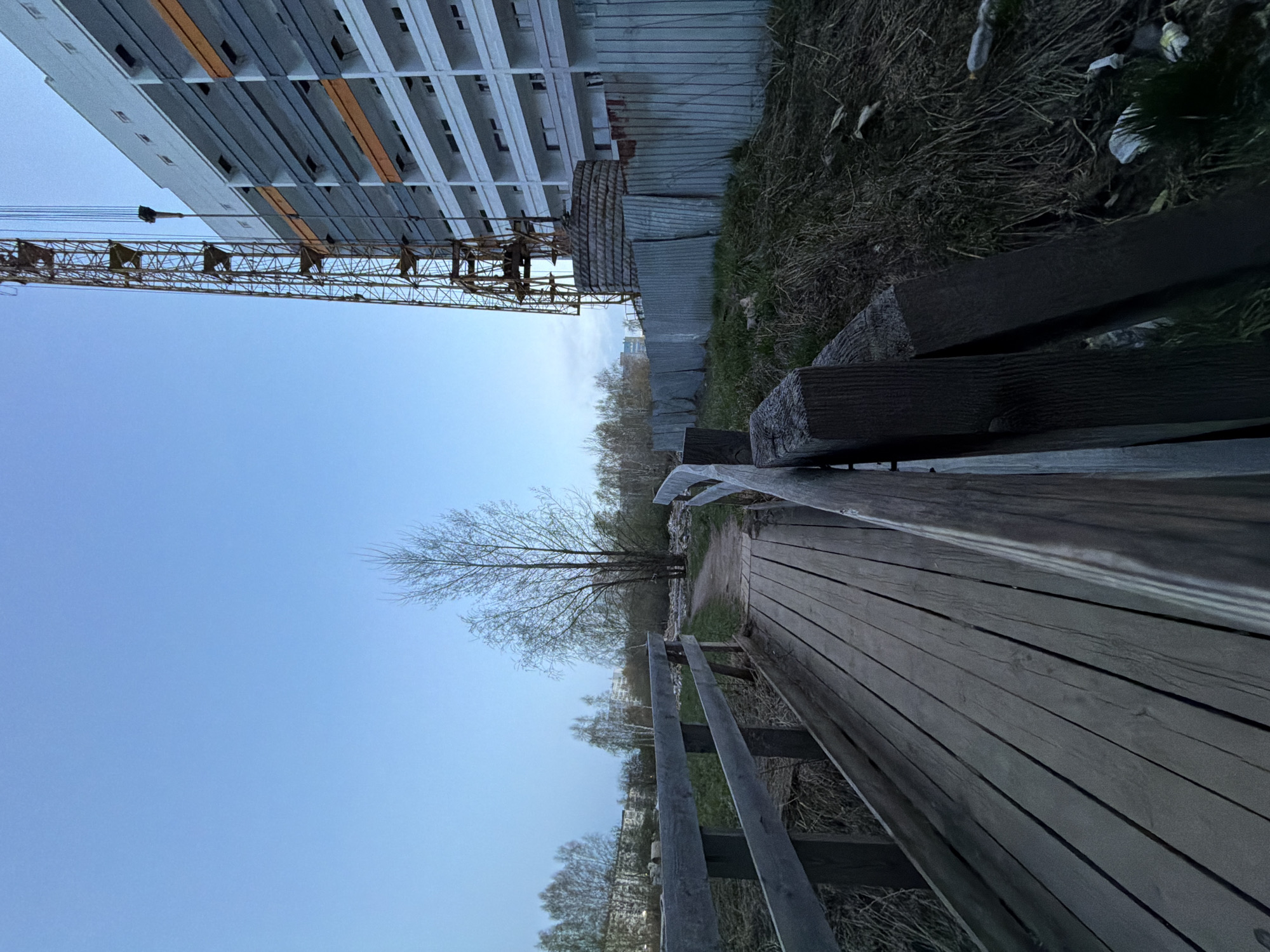The image depicts a view along a wooden boardwalk near a construction site in Chaikovsky, Russia. A tall, unfinished building with a construction crane is visible in the background. A metal fence separates the construction zone from the boardwalk area. The boardwalk has wooden railings. The sky is a clear blue. The scene appears to be captured in the early evening or morning. There are no people visible. Vegetation is present along the edges of the boardwalk and beyond the fence. Litter is also observed near the fence.