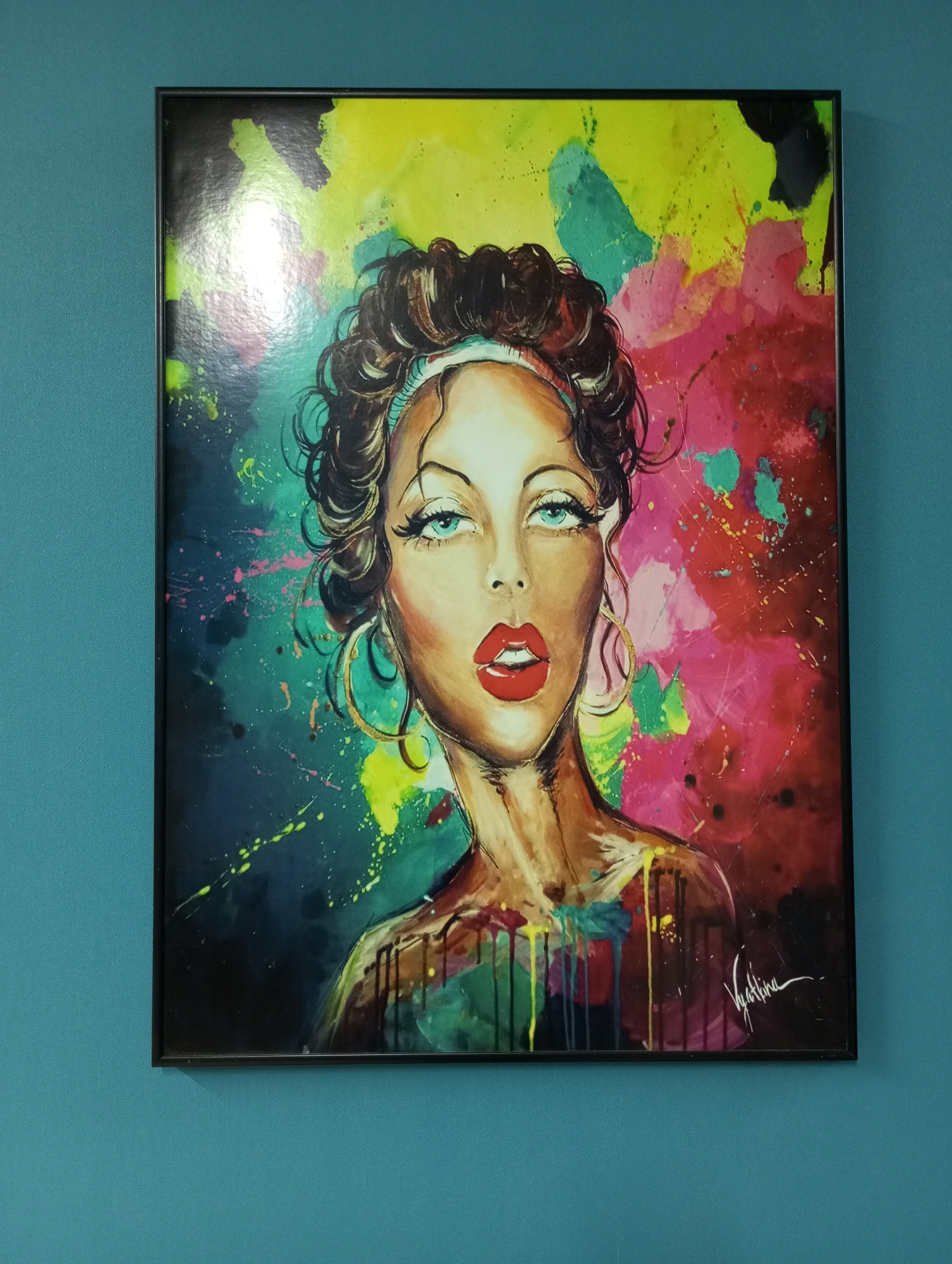 The image shows a framed painting hanging on a teal wall. The painting depicts a woman with curly brown hair, fair skin, red lips, and green eyes. She is wearing gold hoop earrings and a white headband. The background of the painting is a colorful splash of yellow, pink, green, blue, and black. The painting is signed "Vyatkina" in the bottom right corner. There is no information to suggest that the image was taken in the city of Pervomaysky, Russia.