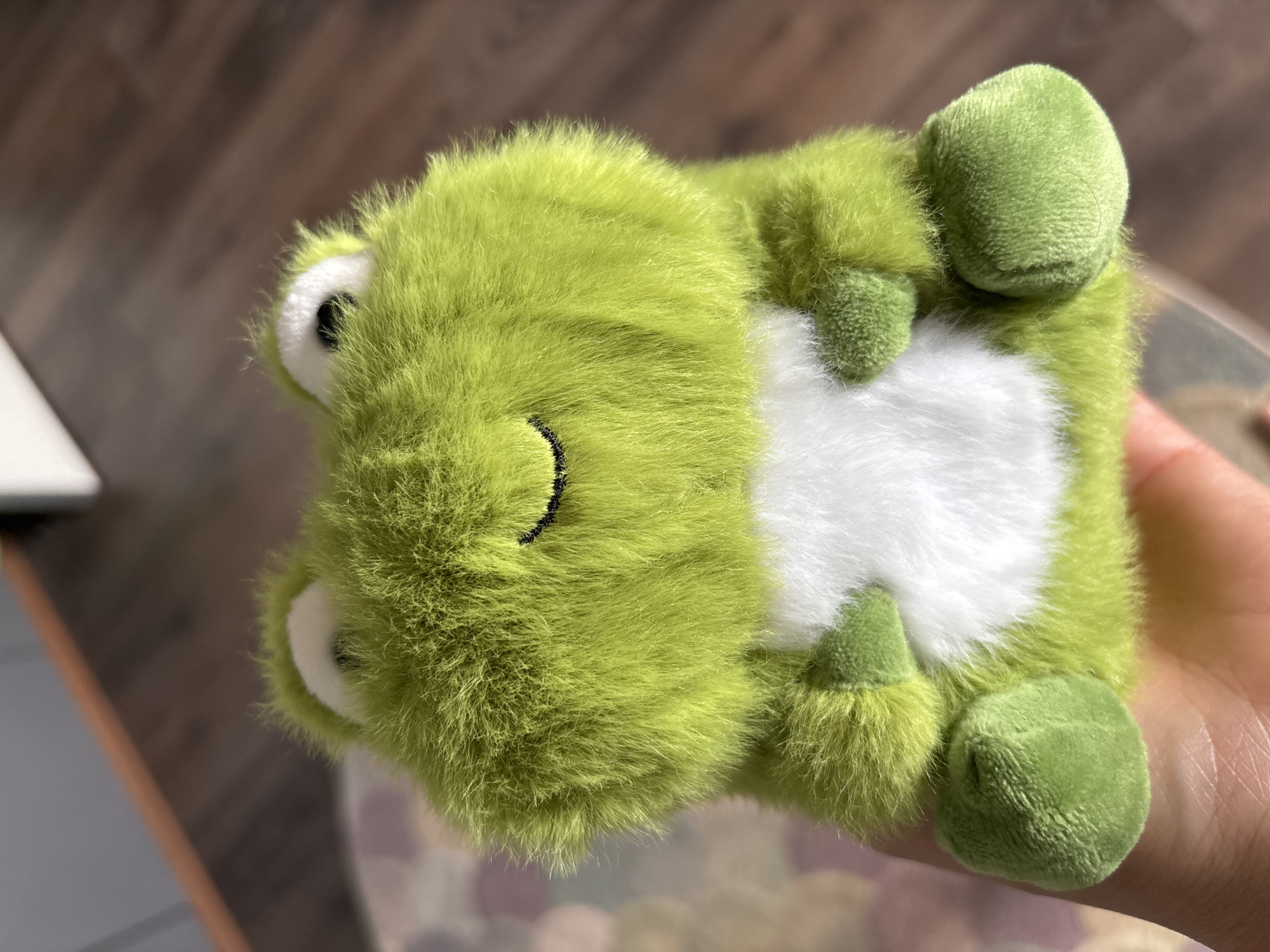 This is a close-up, overhead shot of a small, fuzzy, lime-green stuffed frog held in a person's hand. The frog is positioned on its back, with its four limbs visible. Its belly is white, and its eyes are black with white accents, giving it a cartoonish appearance. A black stitched smile is visible on its face. The background consists of a wooden surface and out-of-focus colorful elements, possibly a patterned blanket or mat. The lighting suggests it is daytime, likely indoors. There is no visible text. The hand holding the frog appears to be a light skin tone. The overall impression is one of a cute, cuddly toy.