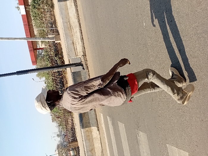 The image portrays a lone individual walking from left to right across a paved road under a clear sky in Sharada, Nigeria. The dark-skinned individual wears a light-colored baseball cap, a visibly worn muted brown or gray long-sleeved shirt with tears, light-colored trousers, and light-colored slip-on shoes, with the left shoe partially disengaged at the heel. A red undergarment is visible at the waistline. A light-colored object is present on the ring finger of the individual's right hand. The road surface features white zebra crossing markings on the left. A long shadow is cast on the road, indicating sunlight from the upper left. To the right of the road, concrete barriers separate the street from a raised area. Beyond these barriers, a dark metal fence borders dense green vegetation. In the background, a structure with a prominent red roof supported by white pillars, consistent with a fuel station canopy, is visible alongside utility poles.
