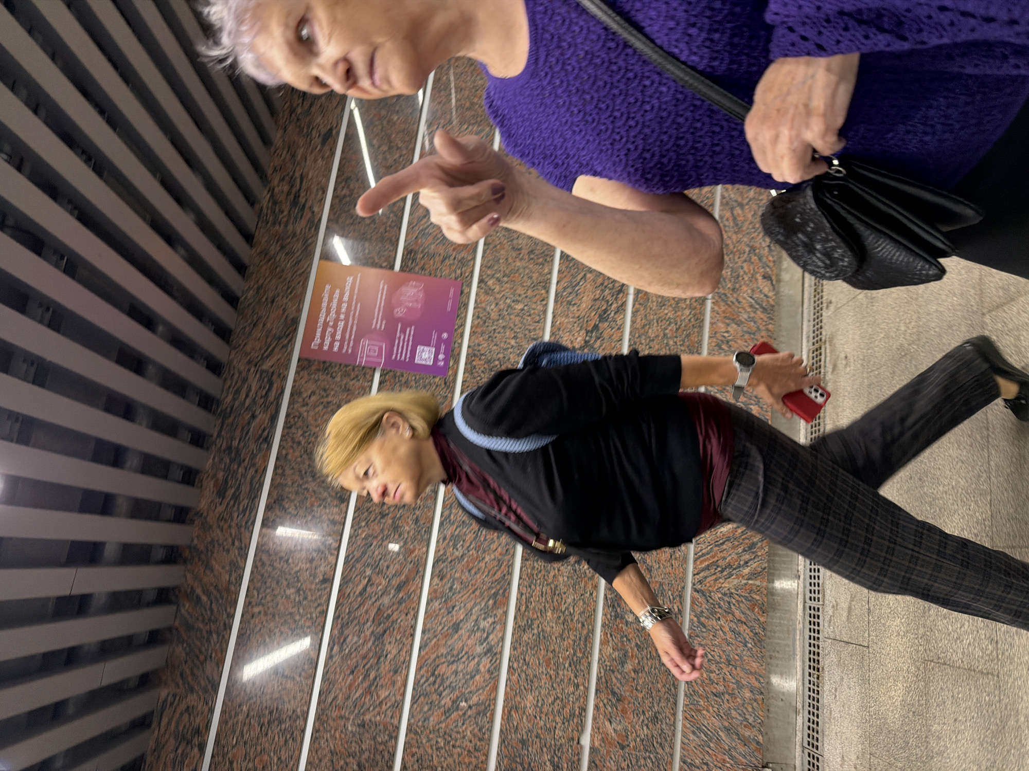 Two women are walking in a subway station. The woman on the left, with blonde hair, is wearing a black sweater, a burgundy top, and plaid pants. She has a watch on her left wrist and is holding a red smartphone in her right hand. The woman on the right, with short grey hair, is wearing a purple textured sweater and is pointing with her right index finger. She has a black handbag. The background features a granite-patterned wall and a slatted ceiling. A sign with Cyrillic text is visible on the wall. The overall impression is that of a public transit environment in Moscow, Russia.