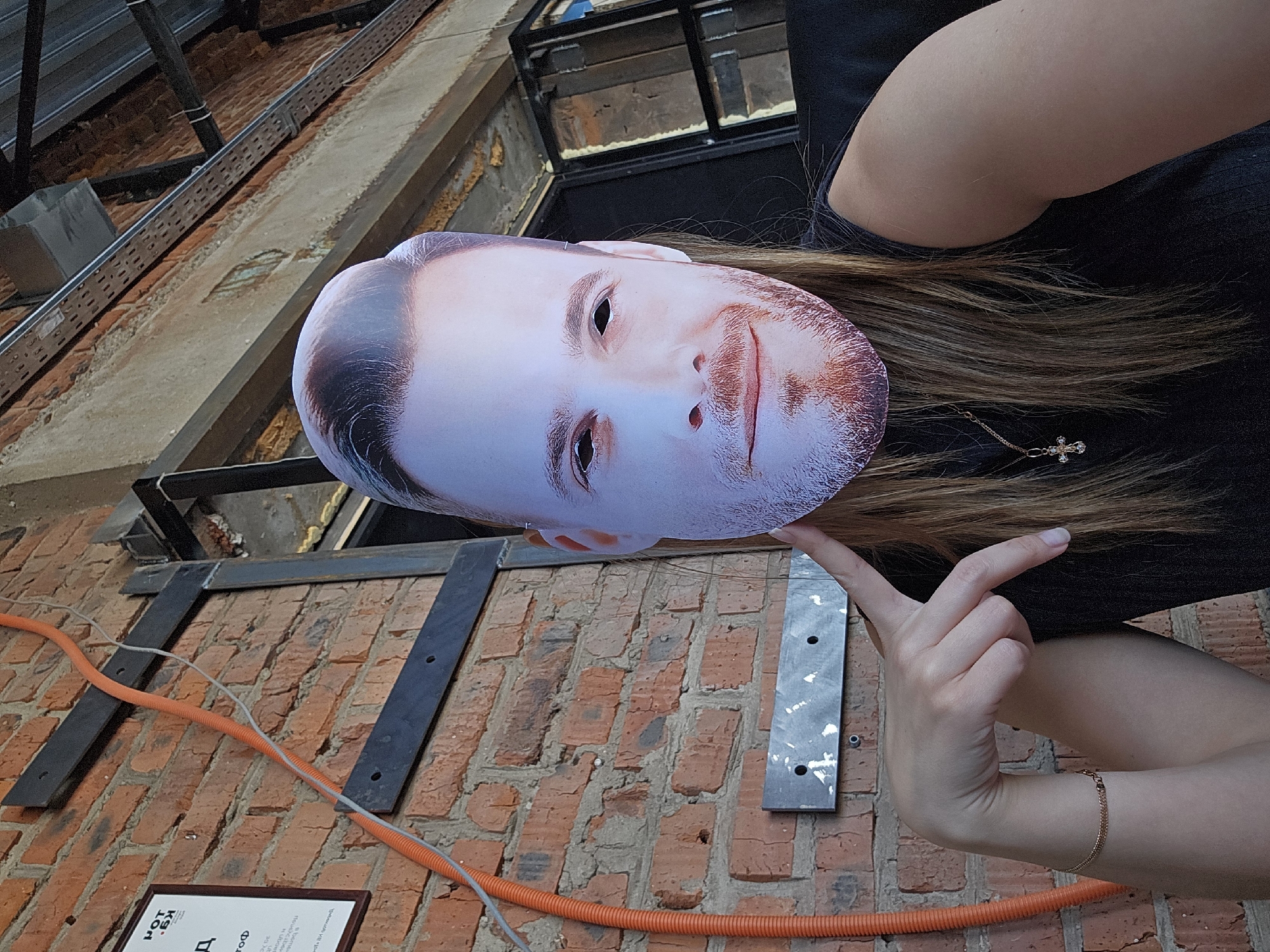A person is holding a face mask of a bearded man in front of them in an indoor location. The background consists of an exposed brick wall with metal supports and orange conduit running along it. There is a framed sign on the wall, partially obscured. The person holding the mask is wearing a black shirt, and has long brown hair. They are also wearing a gold bracelet. The environment appears to be an industrial-style interior with exposed brick and metal infrastructure. The city of Chaikovsky, Russia is not evident in this image.