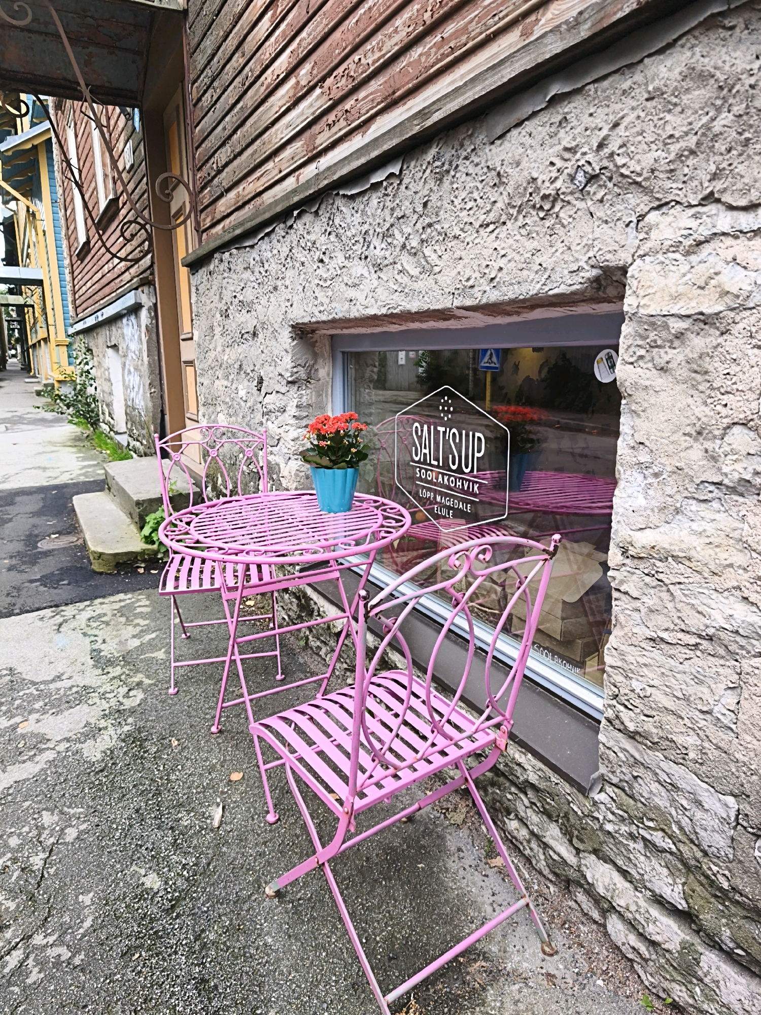 This is a photograph taken in Tallinn, Estonia, of a pink wrought-iron bistro set, consisting of a round table and two chairs, placed on a wet, textured asphalt surface next to a stone building. The building has a rough, grey stone foundation and a wooden facade above it with red-brown, weathered planks. A window is visible in the stone wall, reflecting the scene and displaying a white hexagonal logo with the text "SALT'S UP SOOLAKOHVIK LÕPP MÄGEDALE ELULE". Inside the window, a blue pot with orange flowers is visible on a pink surface, suggesting a seating area within the establishment. The surrounding environment appears to be a narrow, paved walkway or street with some greenery and stone steps to the left, leading towards other buildings with colorful wooden facades. The lighting suggests an overcast or shaded day.