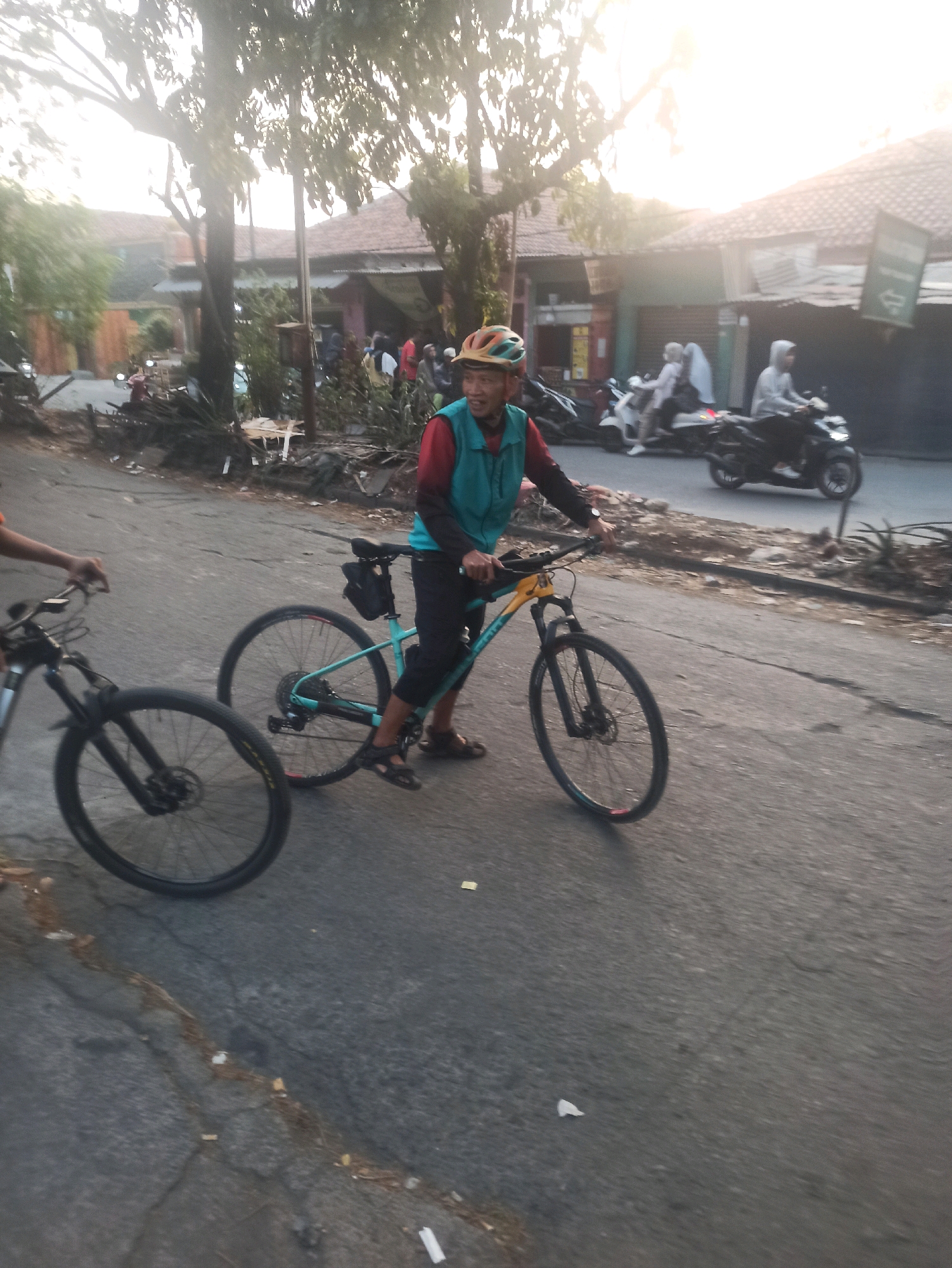In Telagamurni, Indonesia, a man wearing a teal vest over a red shirt and dark pants, along with a helmet, is seen on a teal and yellow bicycle. He is on a paved road. Another bicycle, mostly black, is visible to his left, with its front wheel in the foreground and the rider's arm extended. In the background, there are buildings with tiled roofs, trees, and several motorcycles. Some of the motorcycles appear to be parked, while others have riders. The lighting suggests it is either early morning or late afternoon, with the sun appearing bright and slightly hazy. The road surface shows some cracks and debris.
