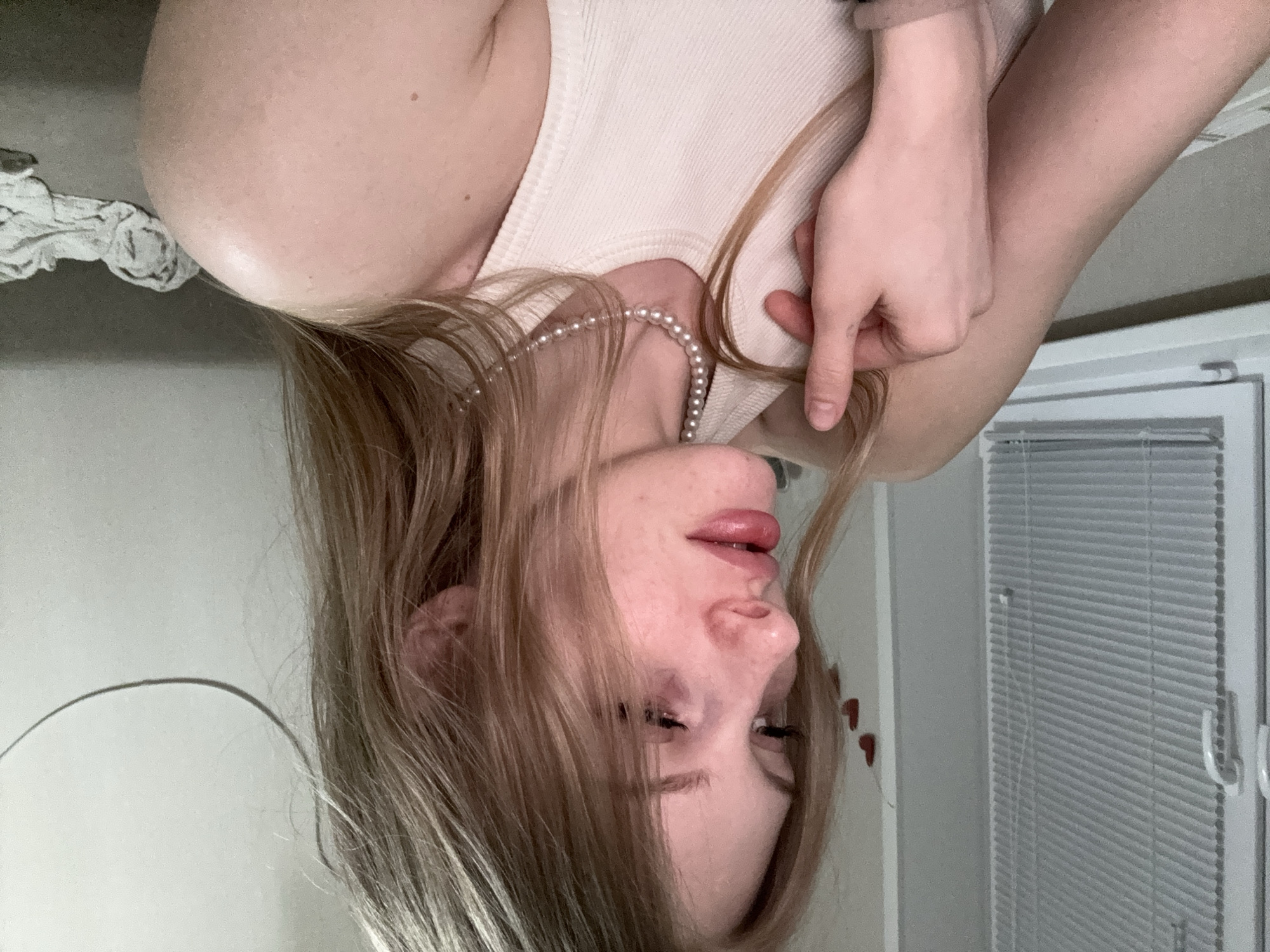 A young woman with blonde hair and fair skin is shown in a close-up, profile view, looking down and to her left. She is wearing a cream-colored tank top and a pearl necklace. Her left hand is resting near her neck. Behind her, a window with blinds is visible on the left, and a sofa with a grey blanket is in the background on the right. The lighting suggests an indoor setting, possibly a residential room, and there are some small red heart-shaped decorations on the wall behind her. No specific location in the city of Perm, Russia can be determined from the image.