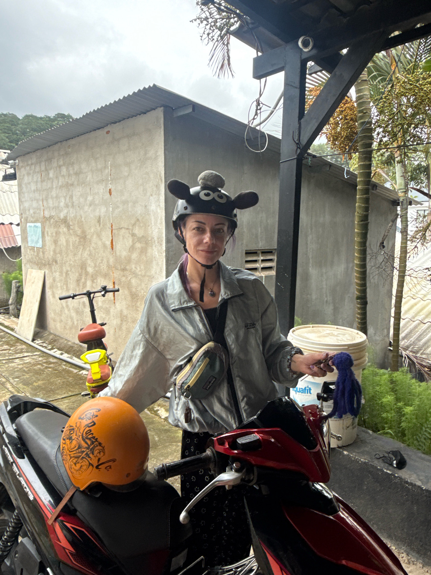 In Hiệp Thạnh, Vietnam, a woman stands in front of a motorcycle. She is wearing a silver jacket over a black top, black patterned pants, and a helmet with cartoonish black ears. A crossbody bag is slung over her left shoulder. In her right hand, she holds a small, fuzzy purple object. To her left, an orange helmet with a black design rests on the motorcycle seat. Behind her, a pale concrete building with a corrugated metal roof is visible, along with a small bicycle. The ground appears wet, suggesting recent rain. A large white bucket stands to her right, with lush green foliage in the background. A palm tree with developing fruit is also present. A security camera is mounted on a dark wooden beam above the woman.