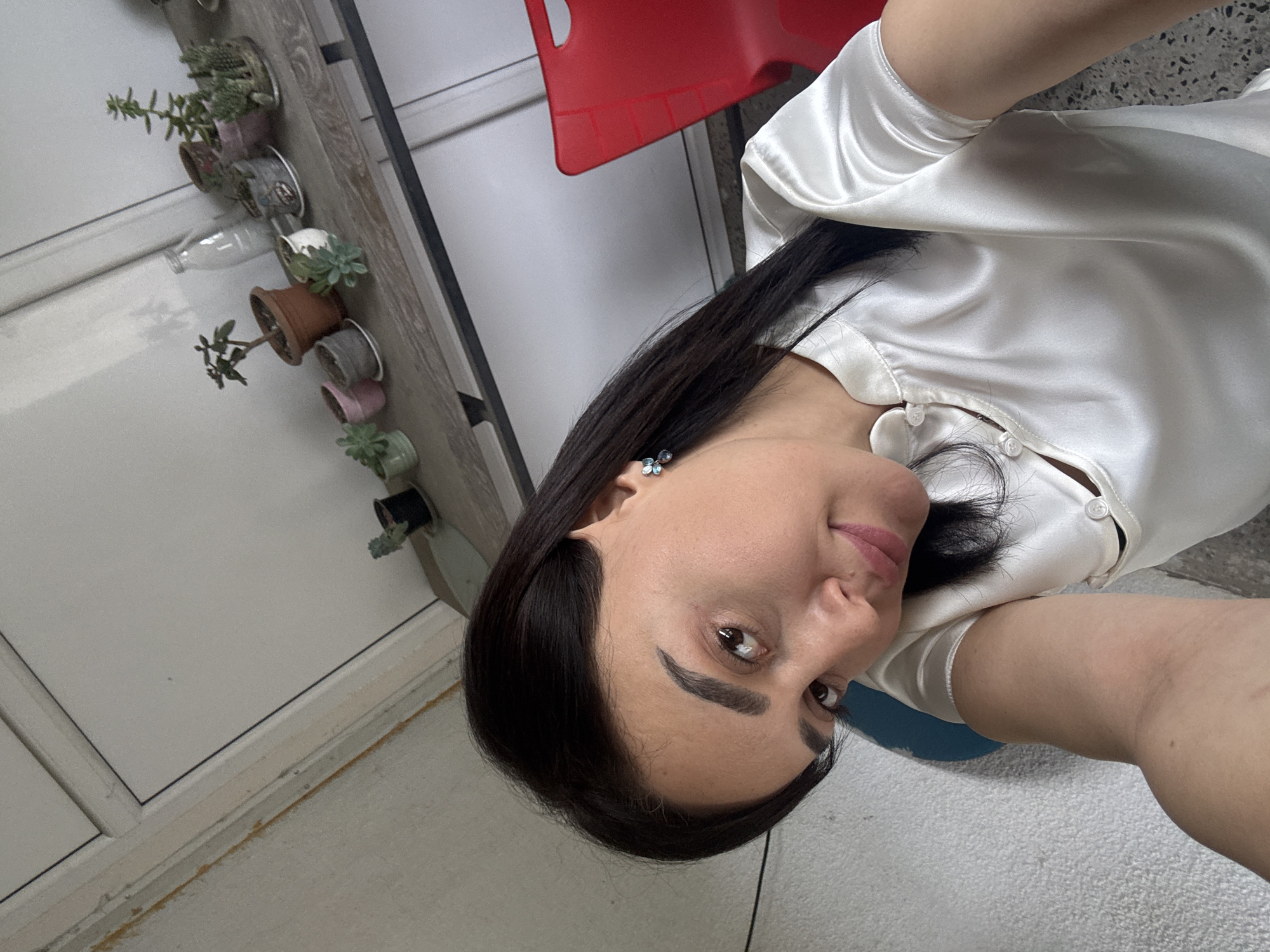 A dark-haired woman is positioned in the foreground, wearing a white, button-up shirt. She is indoors, seated on a blue object, which is likely a chair or stool. Behind her is a red chair. A series of potted plants are arranged along a shelf or ledge. A white door is also visible. While no definitive information indicates the location as Tbilisi, Georgia, that is potentially the case.