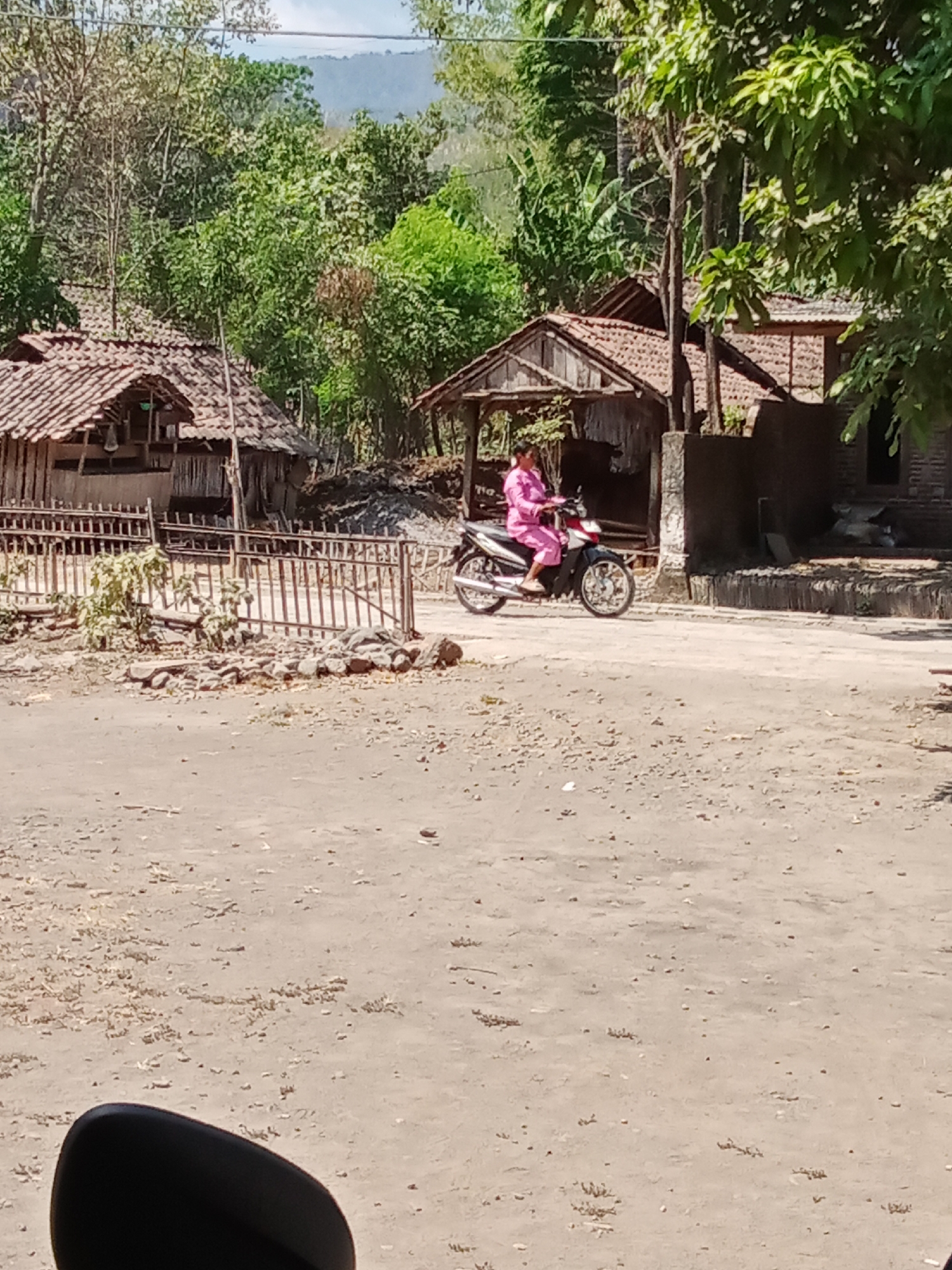In Selowogo, Indonesia, a woman wearing a bright pink outfit rides a motorcycle on a dirt road. The road is lined with a rustic metal fence and a few scattered rocks. Behind the fence, traditional Indonesian houses with tiled roofs are visible, surrounded by lush green trees and vegetation. The sky above is a clear blue. In the foreground, part of a black motorcycle mirror is visible, indicating the viewpoint of the observer.