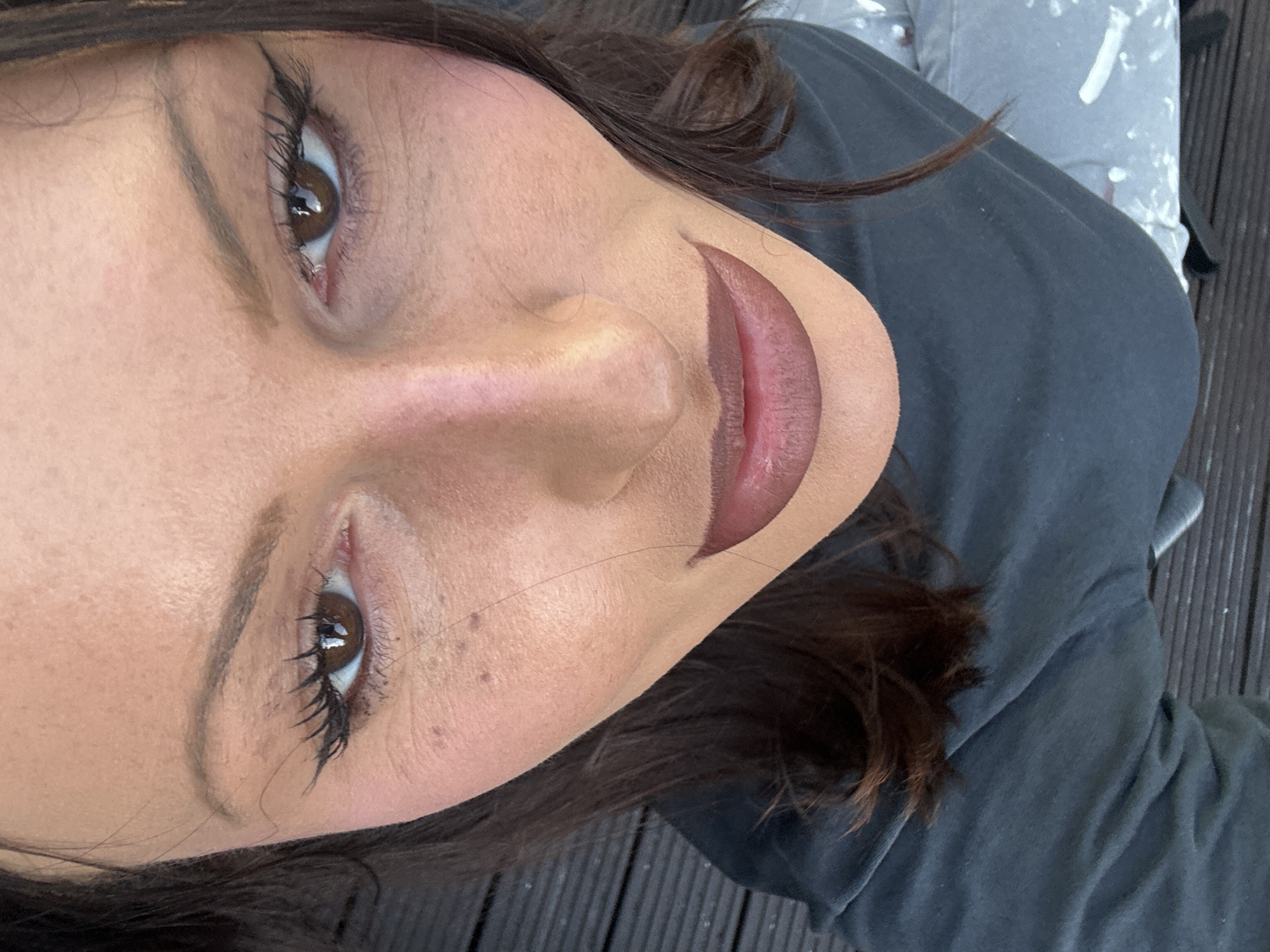 A close-up, tilted portrait captures a woman with brown hair and fair skin. Her makeup includes winged eyeliner and lip liner. She appears to be wearing a dark gray t-shirt. The background features a wooden surface with vertical grooves. There is no indication of being in London, United Kingdom.